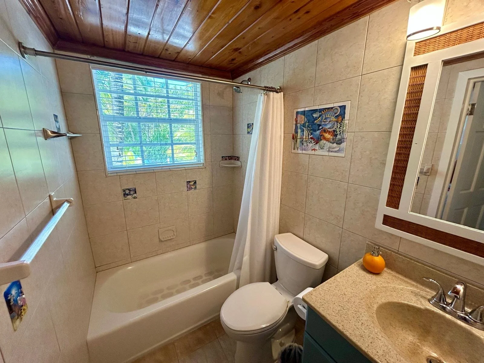 A small bathroom with a bathtub, toilet, and a vanity sink. There is a window with blinds letting in natural light, and a wooden ceiling. The walls are tiled with decorative tiles, and a mirror is above the sink.