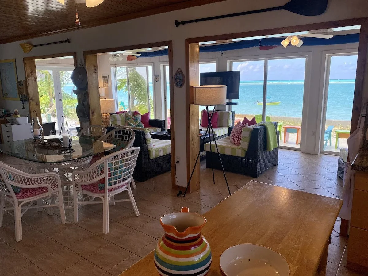 Interior of a beachside living room with sliding glass doors showing the ocean view, wicker furniture with colorful cushions, a dining table with chairs, and tropical decor.