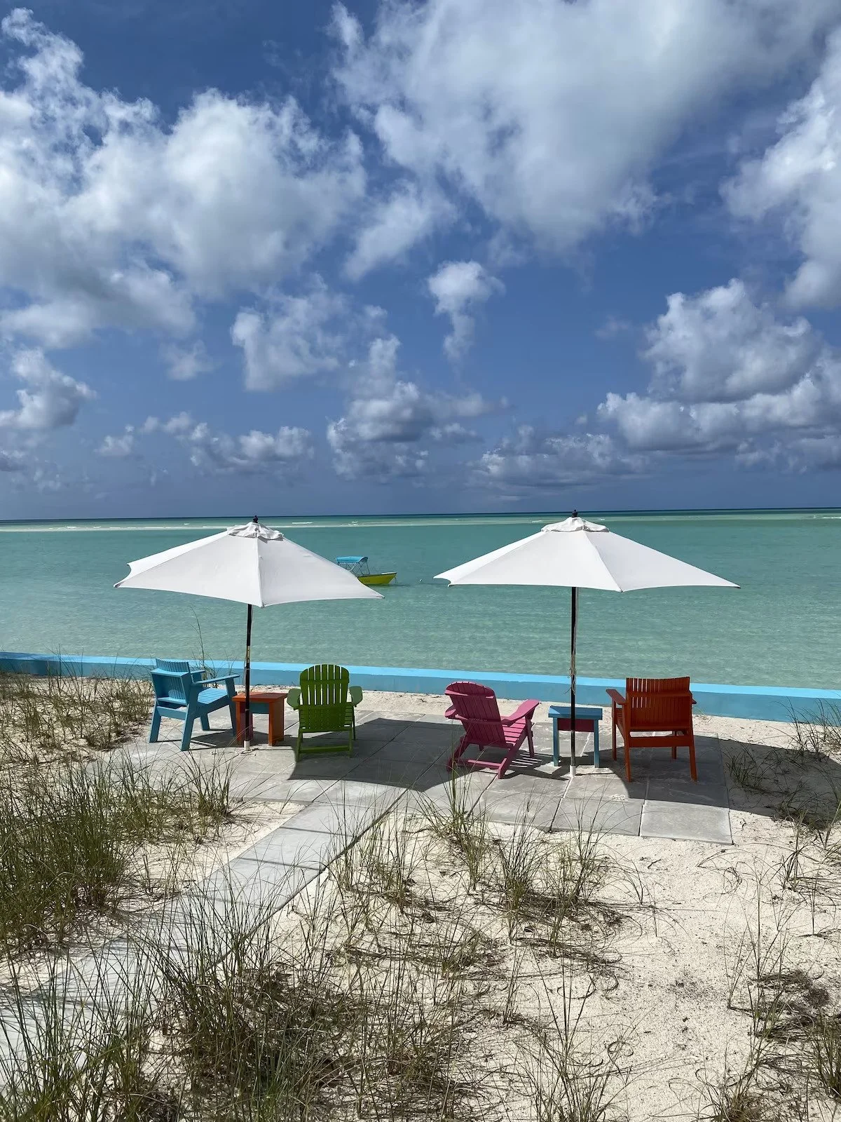 Two white beach umbrellas, four colorful chairs, and a small table on a sandy beach near turquoise water with a cloudy sky.