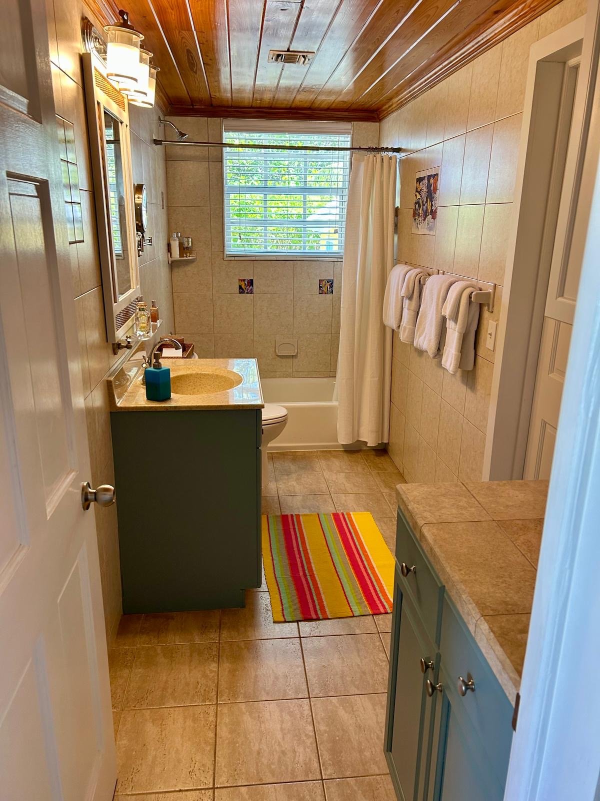 A bathroom with a blue vanity, tan countertops, beige tiled walls and floor, a window with blinds, a shower curtain, and colorful striped rug.