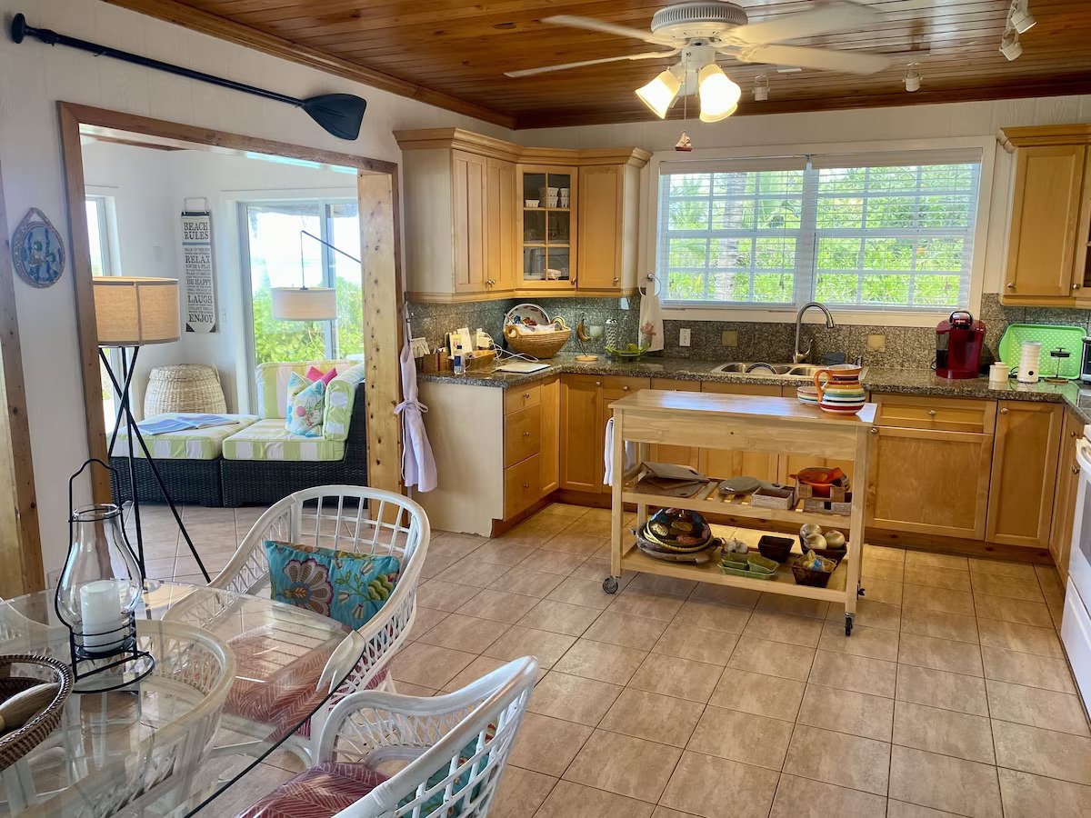 Kitchen with wooden cabinets, granite countertops, and a large window. There is a kitchen island on wheels with dishes and bowls. A dining table with wicker chairs and a candle lantern is in the foreground. A sunlit sitting area with a striped couch 