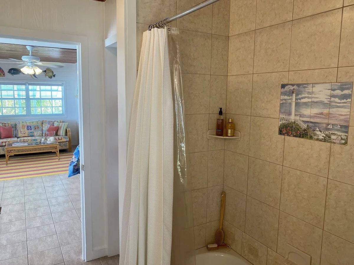 Bathroom shower with beige tiled walls, shower curtain, soap bottles, and a wall art depicting a lighthouse scene.