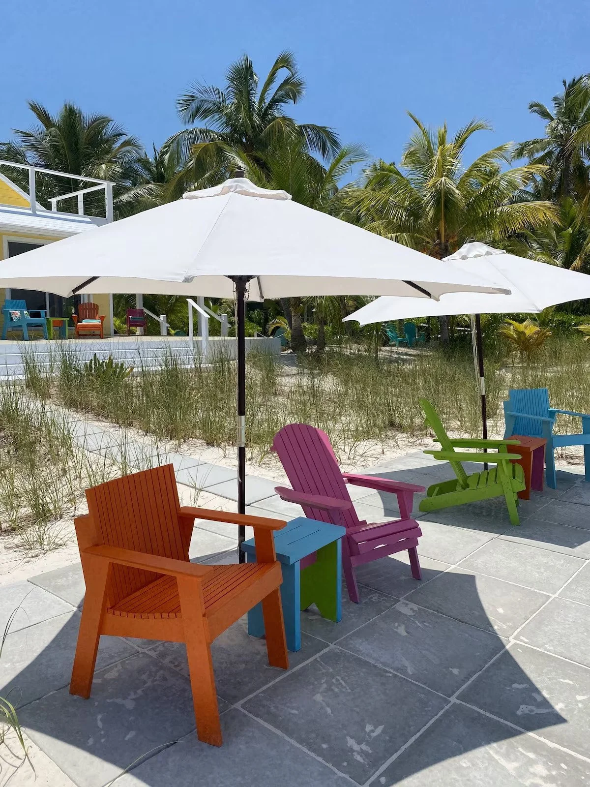 Colorful outdoor chairs and tables with umbrellas on a beachside patio, with palm trees and sand in the background.