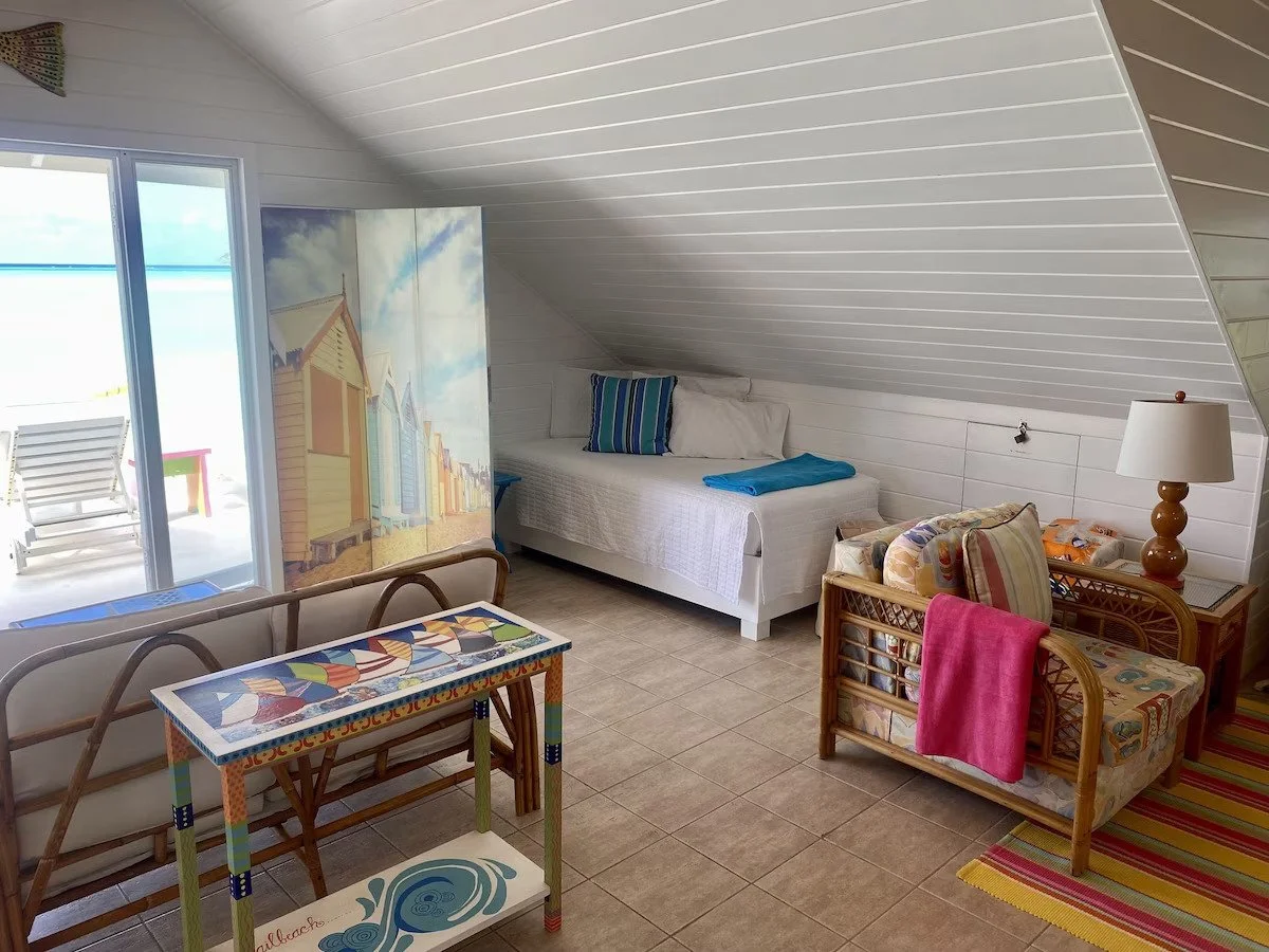 Beach-themed room with a white bed, blue pillow, colorful armchair, wooden side table with a lamp, small table with a boat-themed design, sliding glass door leading to a beach with chairs, and a folding screen with beach huts painted on it.