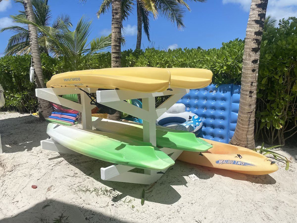 Stacked kayaks and paddleboards on sandy beach with palm trees and blue sky in the background.