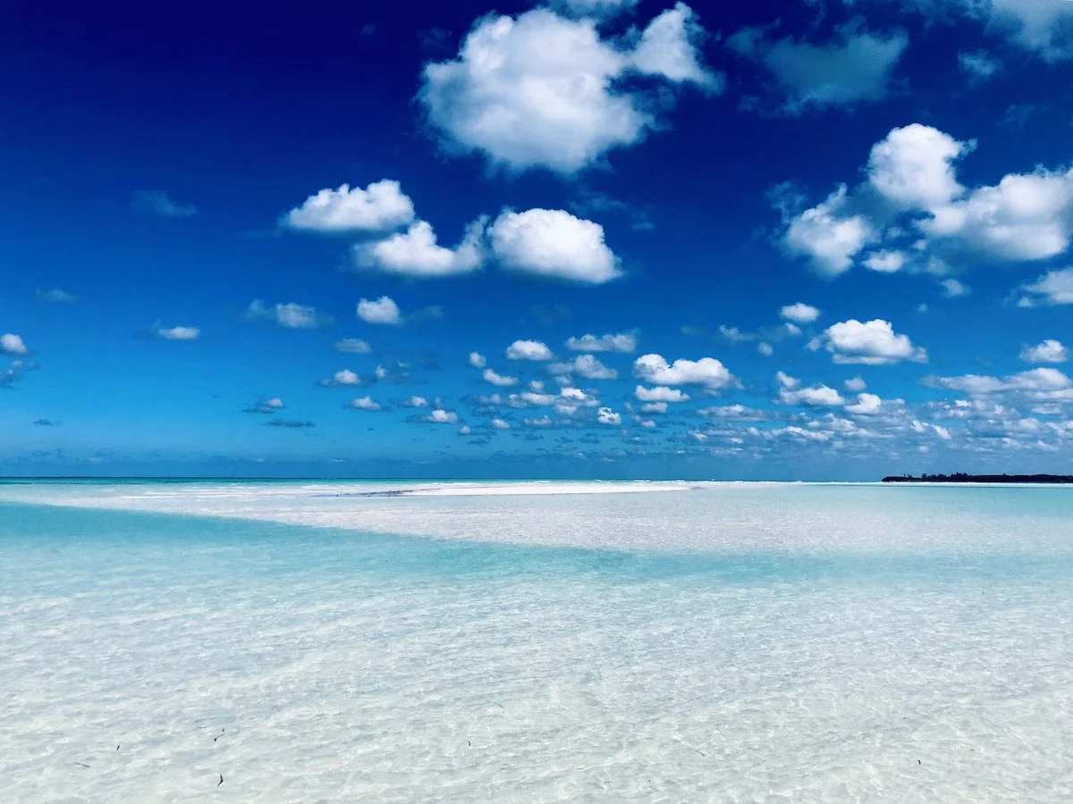A serene beach with white sand, turquoise water, and a clear blue sky filled with scattered white clouds.