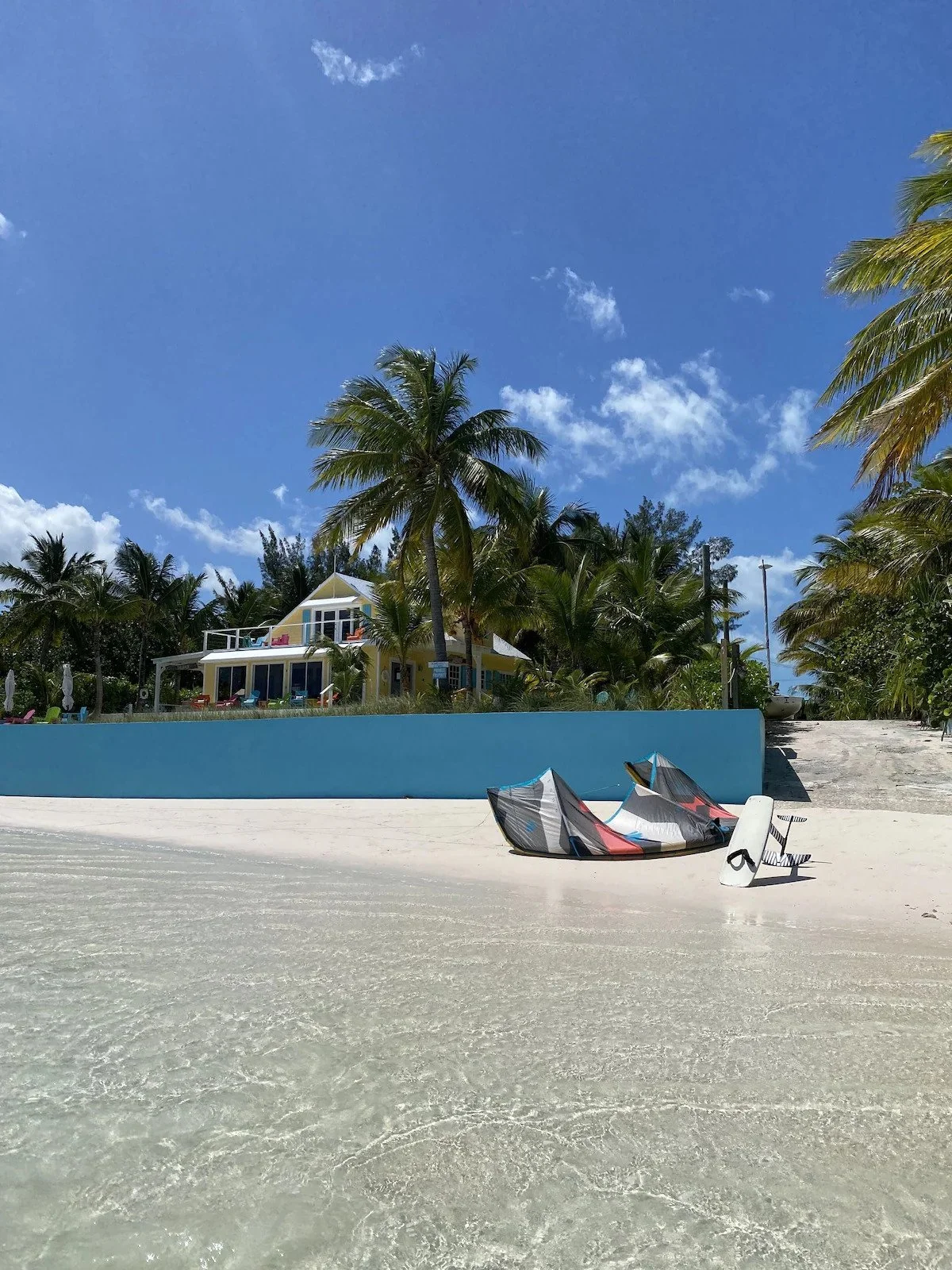 Beach with clear sand, two kites or small sailboat on the ground, a blue wall, palm trees, a yellow house with a balcony, and a partly cloudy blue sky.