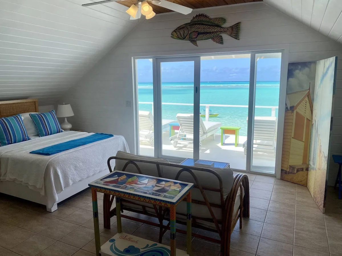 Beachside hotel room with a bed, a small table, a sofa, and a view of the ocean through sliding glass doors, with lounge chairs on the balcony.