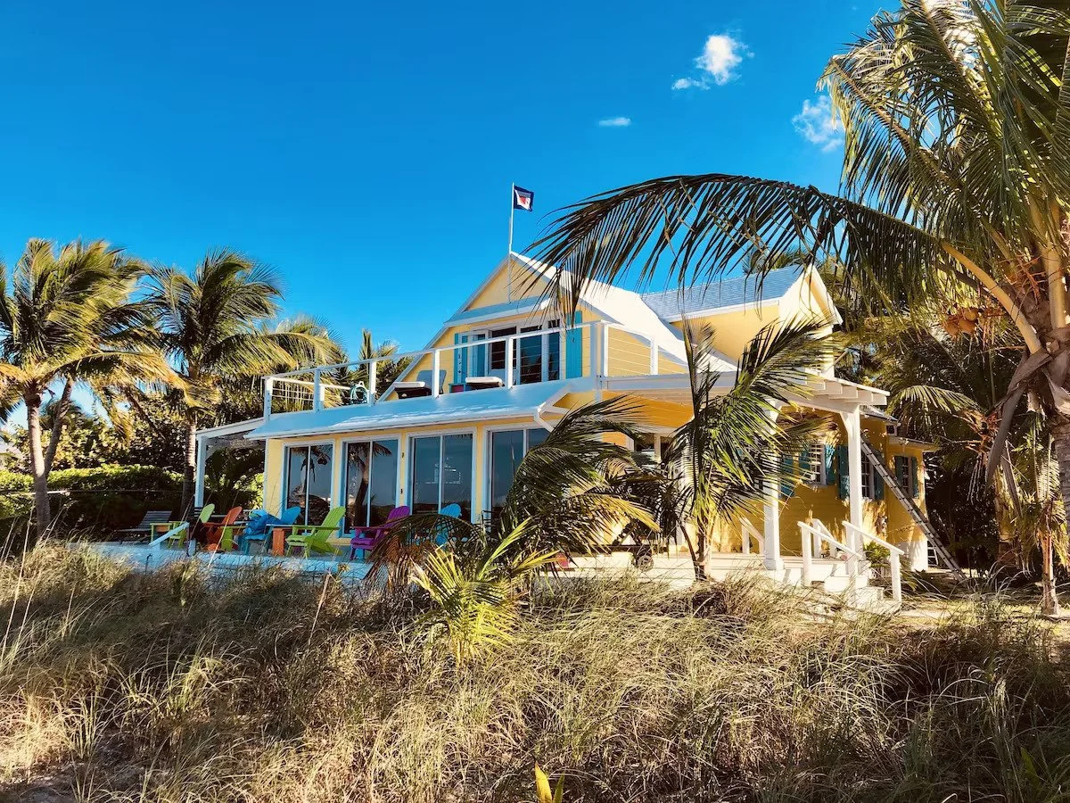 Bright yellow beach house with large glass windows and a wrap-around porch, surrounded by palm trees and tropical plants on a sunny day.