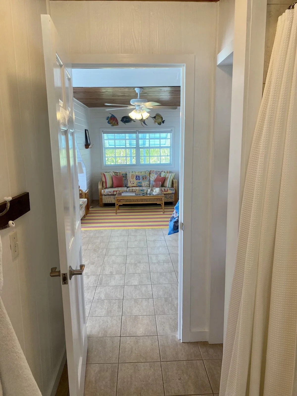 View through doorway into a tropical-themed living room with a rattan sofa, colorful striped cushions, a coffee table, and a window with blinds. Decor includes fish wall art and a ceiling fan with lights.