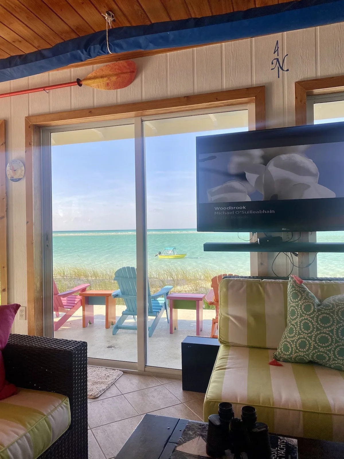 Beach view through sliding glass door with colorful outdoor chairs, boat on water, and TV inside showing the movie 'Woodbrook'.