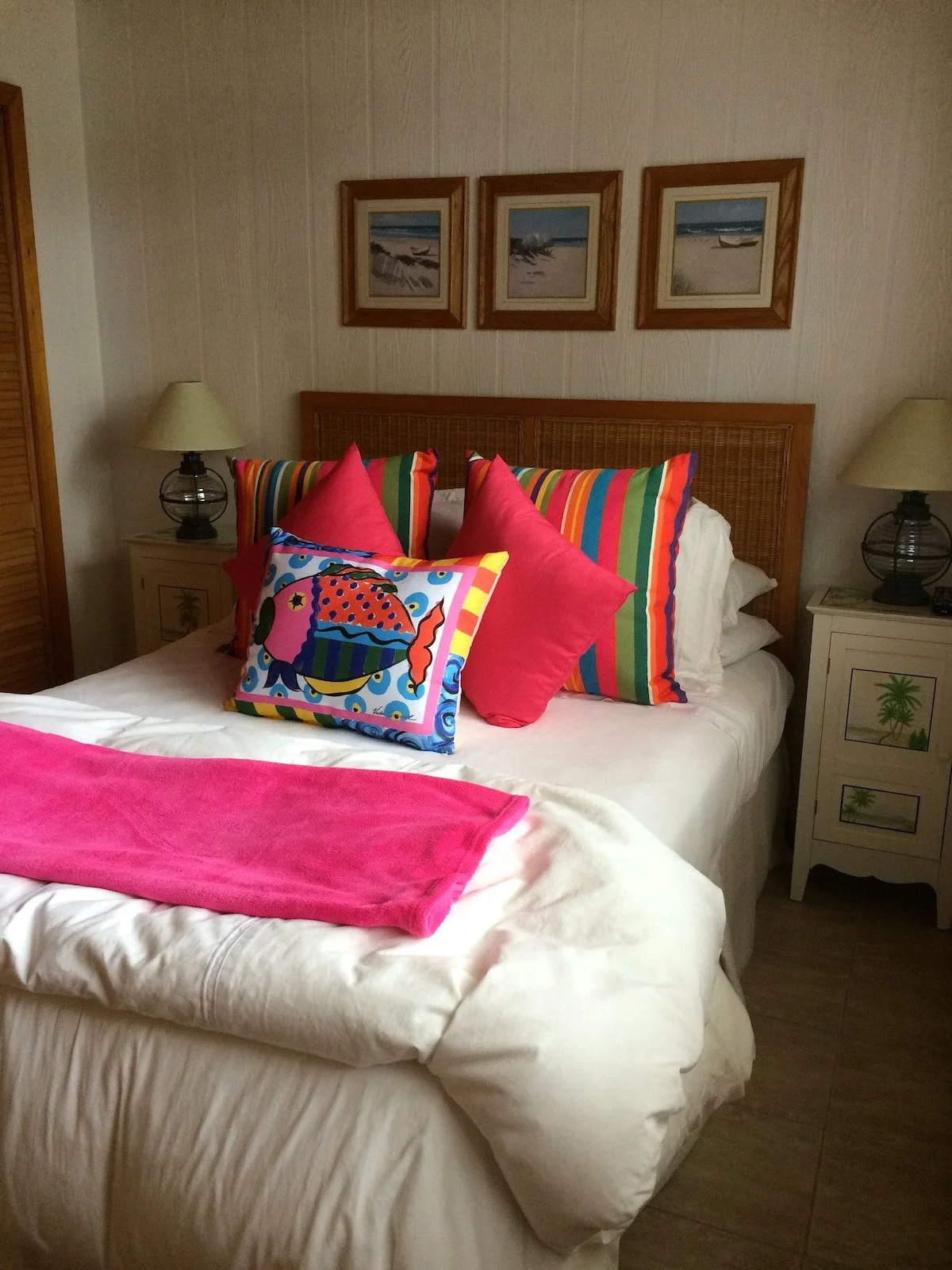 A neatly made bed with white sheets, topped with colorful pillows featuring beach and ocean themes, and with framed beach pictures hanging on the wall behind in a coastal style room.