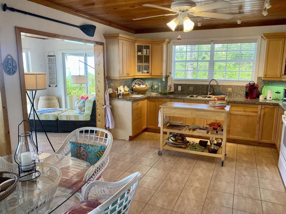 Kitchen with wooden cabinets, granite countertops, a window above the sink, a rolling kitchen island with plates and bowls, and a view into a sunlit living room with a rattan sofa, striped cushions, and a large window.