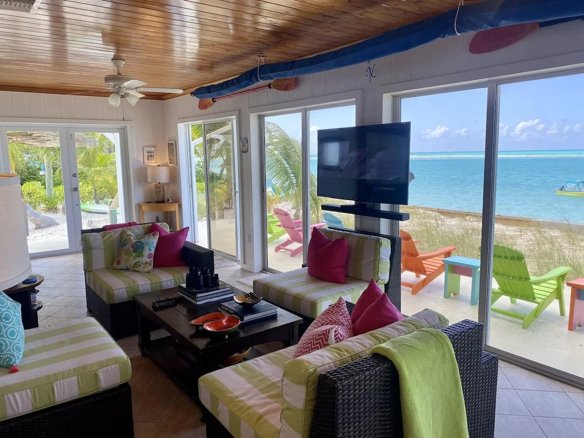 Living room with large sliding glass doors overlooking a beach and ocean, colorful chairs outside, and tropical plants visible through the windows.