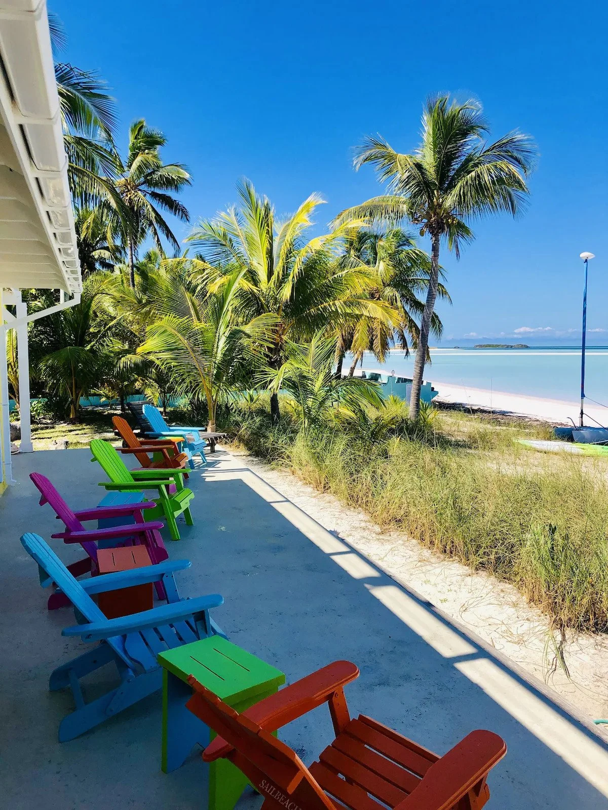Colorful Adirondack chairs on a porch overlooking a beach with palm trees and blue sky.