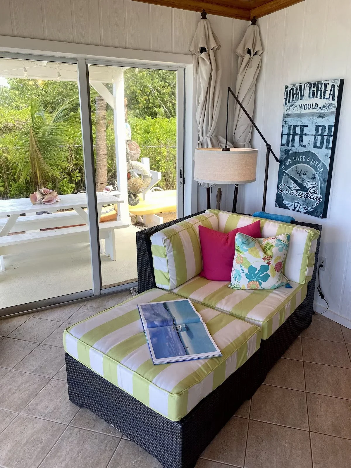 A living room with a colorful striped lounge chair with pillows in pink, green, and floral patterns, positioned near a sliding glass door leading to an outdoor patio. The patio has a white table, chairs, and lush greenery. Inside, a wall-mounted lamp