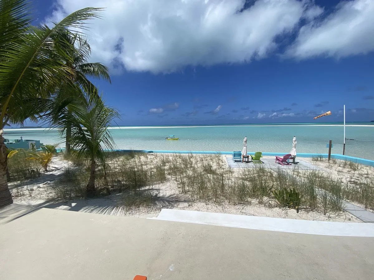 A tropical beach scene with palm trees, sandy shore, lounge chairs, umbrellas, and calm turquoise water under a partly cloudy sky.