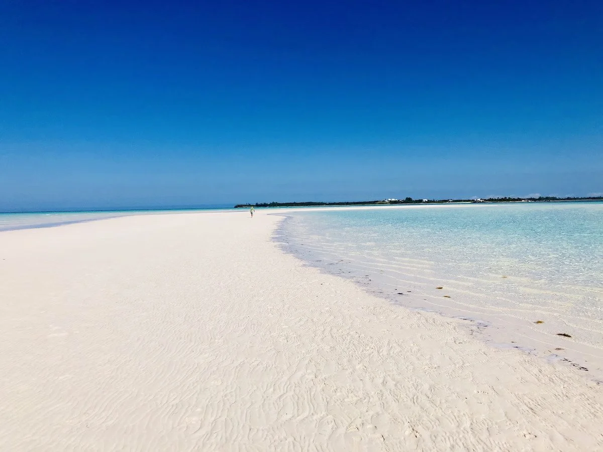 White sandy beach with clear blue water and bright blue sky, distant small boat or structure at the horizon, few people walking along the shoreline.