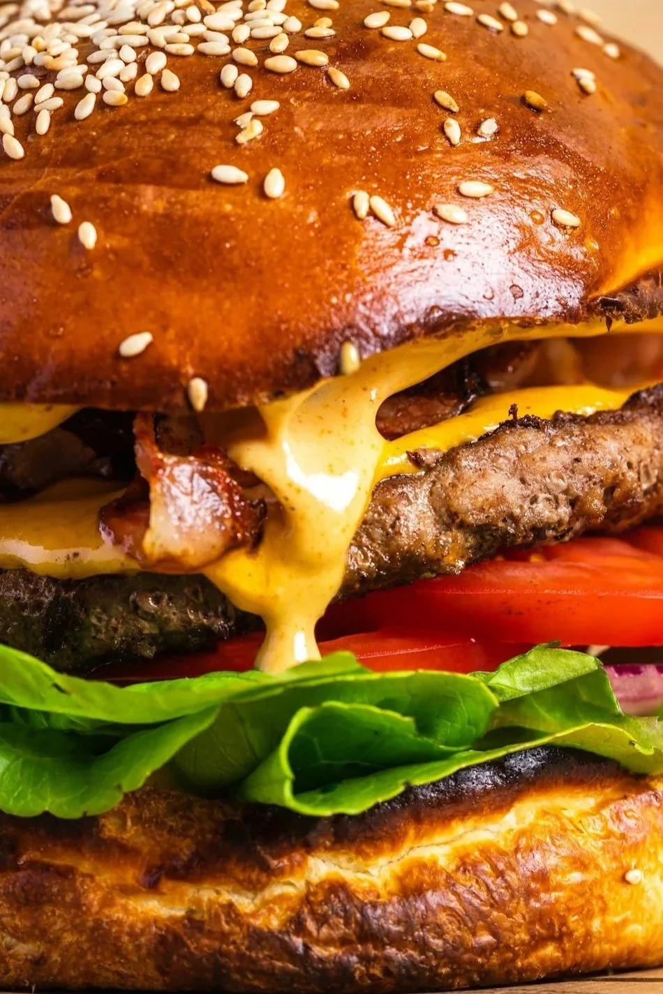 Close-up of a cheeseburger with sesame seed bun, melted cheese, lettuce, tomato, bacon, and a beef patty.