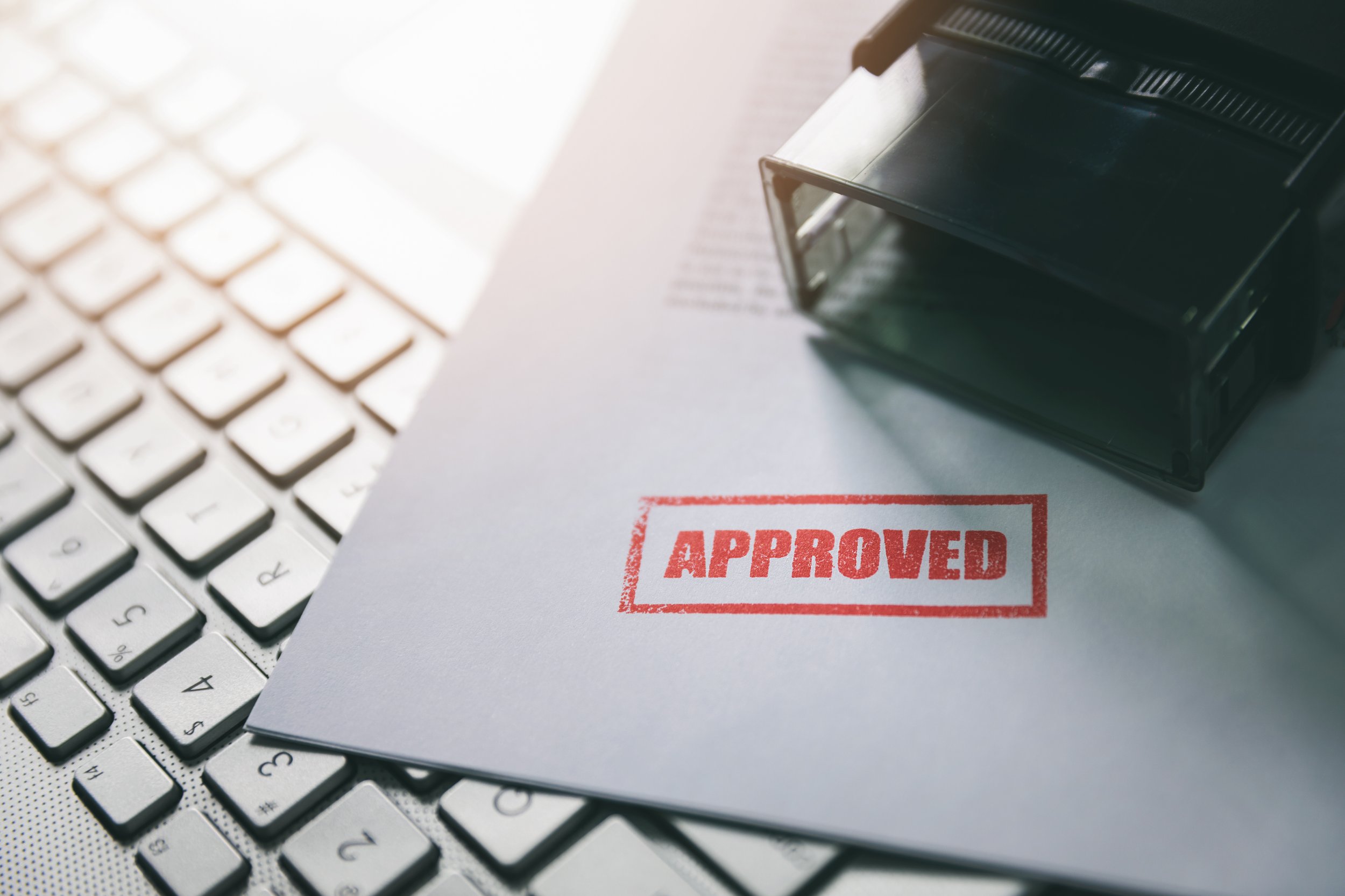 Close-up of a computer keyboard, some paperwork, and a stamp with the word 'APPROVED' on it.
