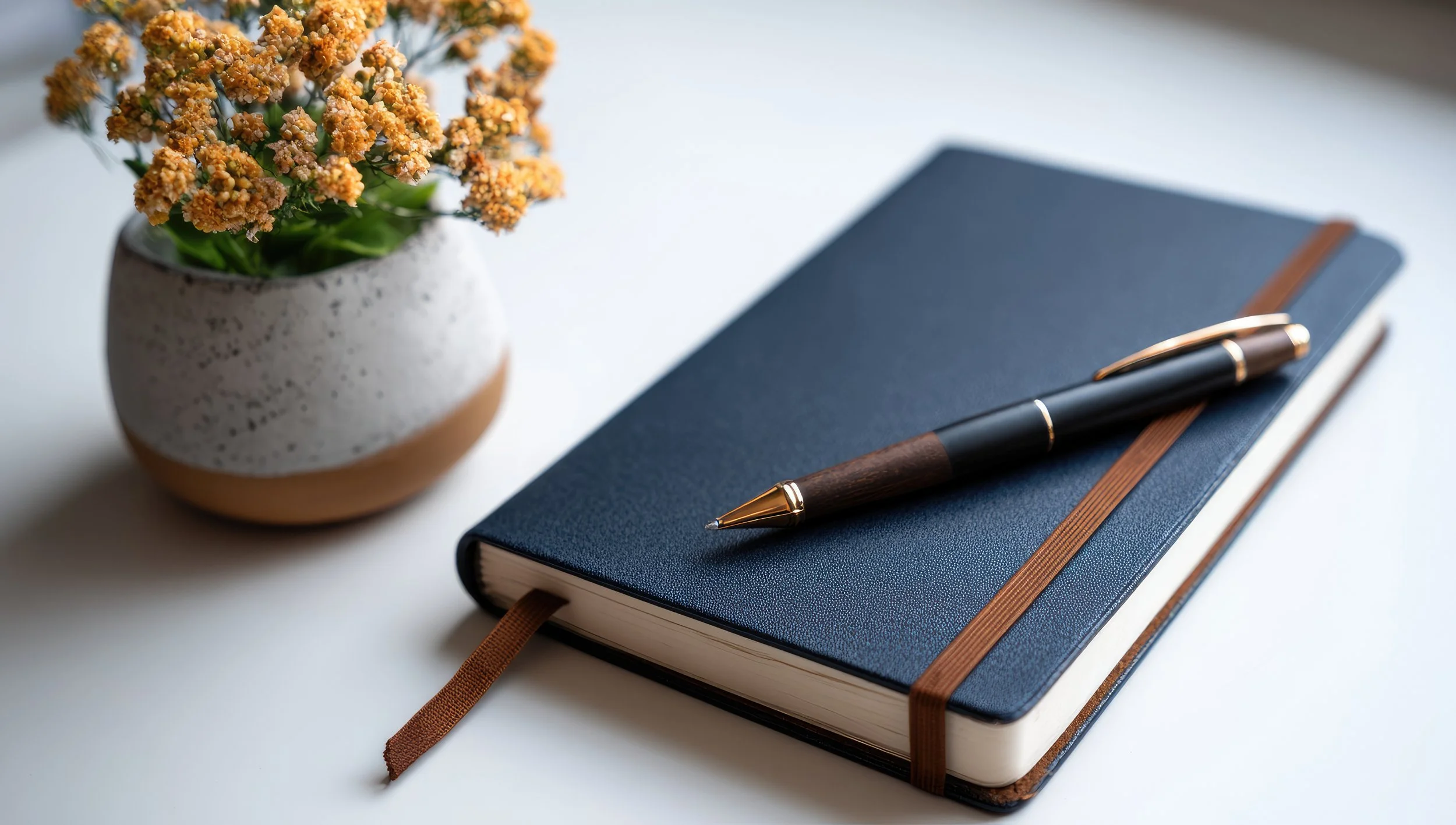 A blue hardcover notebook with an elastic band and a brown ribbon bookmark, accompanied by a black and gold pen on top, placed next to a white and brown speckled ceramic vase with orange flowers, all on a white surface.