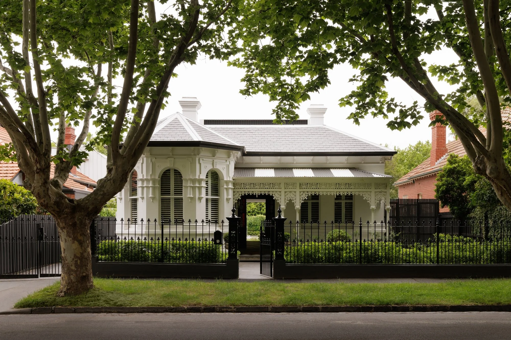 A white Victorian-style house with intricate trim, bay windows, and a black gate, seen from the sidewalk with a large leafy tree in the foreground.