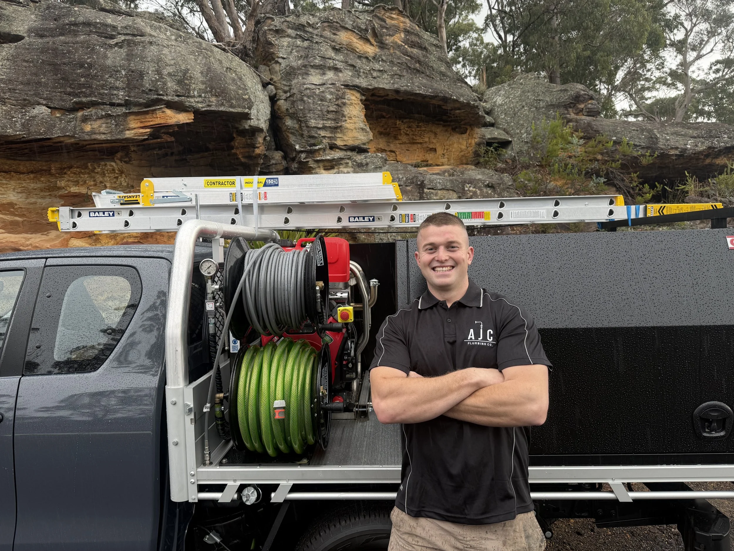 Smiling man in black polo shirt standing with arms crossed in front of work truck with ladder and hose, outdoors near rocky hillside and trees