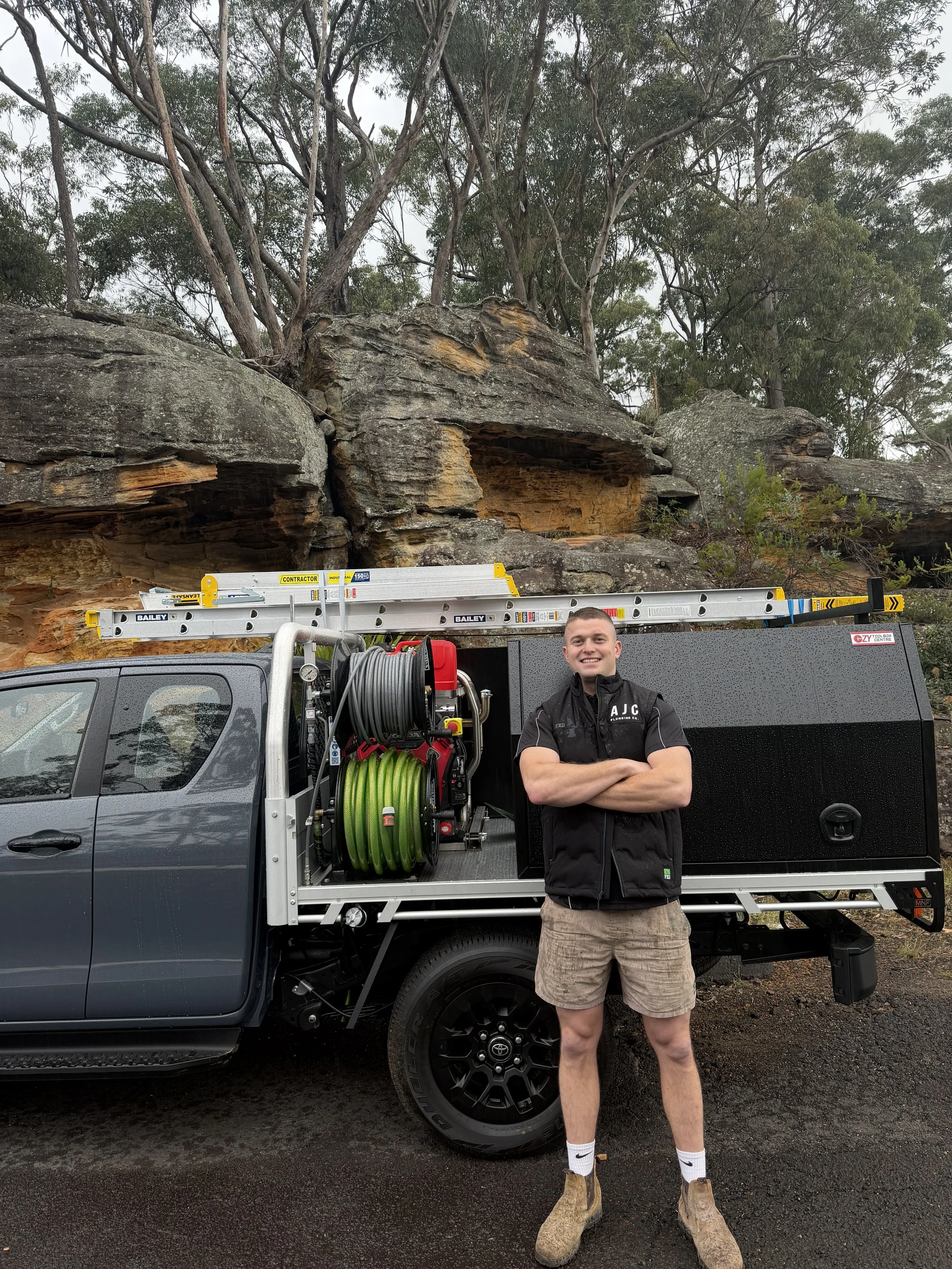 A man standing in front of a black work truck with various tools and equipment, behind him a rocky hillside with trees.