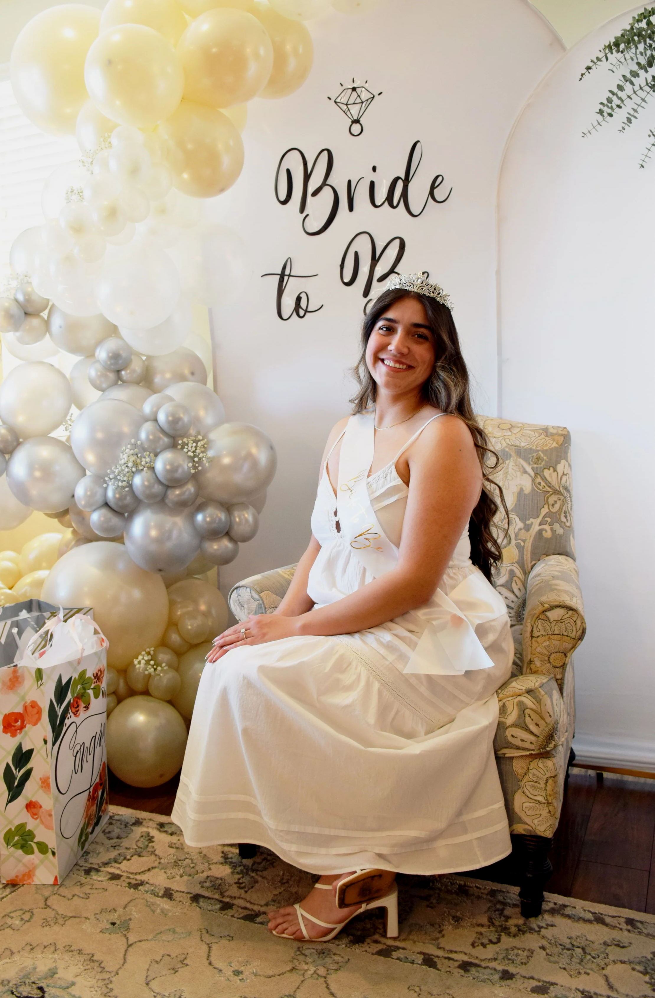 A young woman wearing a crown and white dress sitting on a floral armchair at a bridal shower. She is smiling, with a balloon and gift bag nearby, and a sign in the background reads 'Bride to Be'.