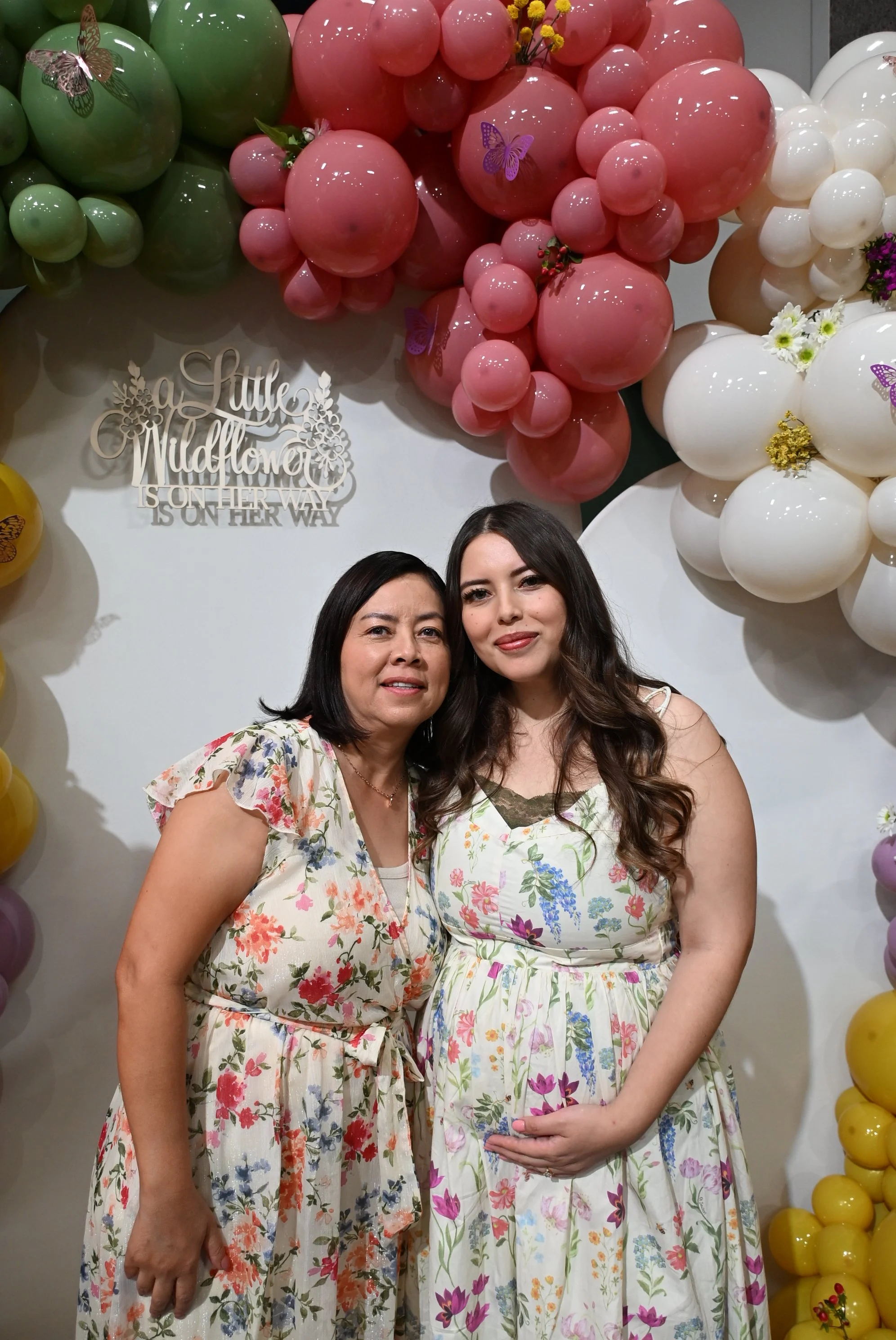Two women standing in front of a colorful balloon decoration with pink, green, white, and yellow balloons and butterfly stickers, at a celebration. One woman is pregnant, wearing a white floral dress, and the other woman is wearing a floral top.