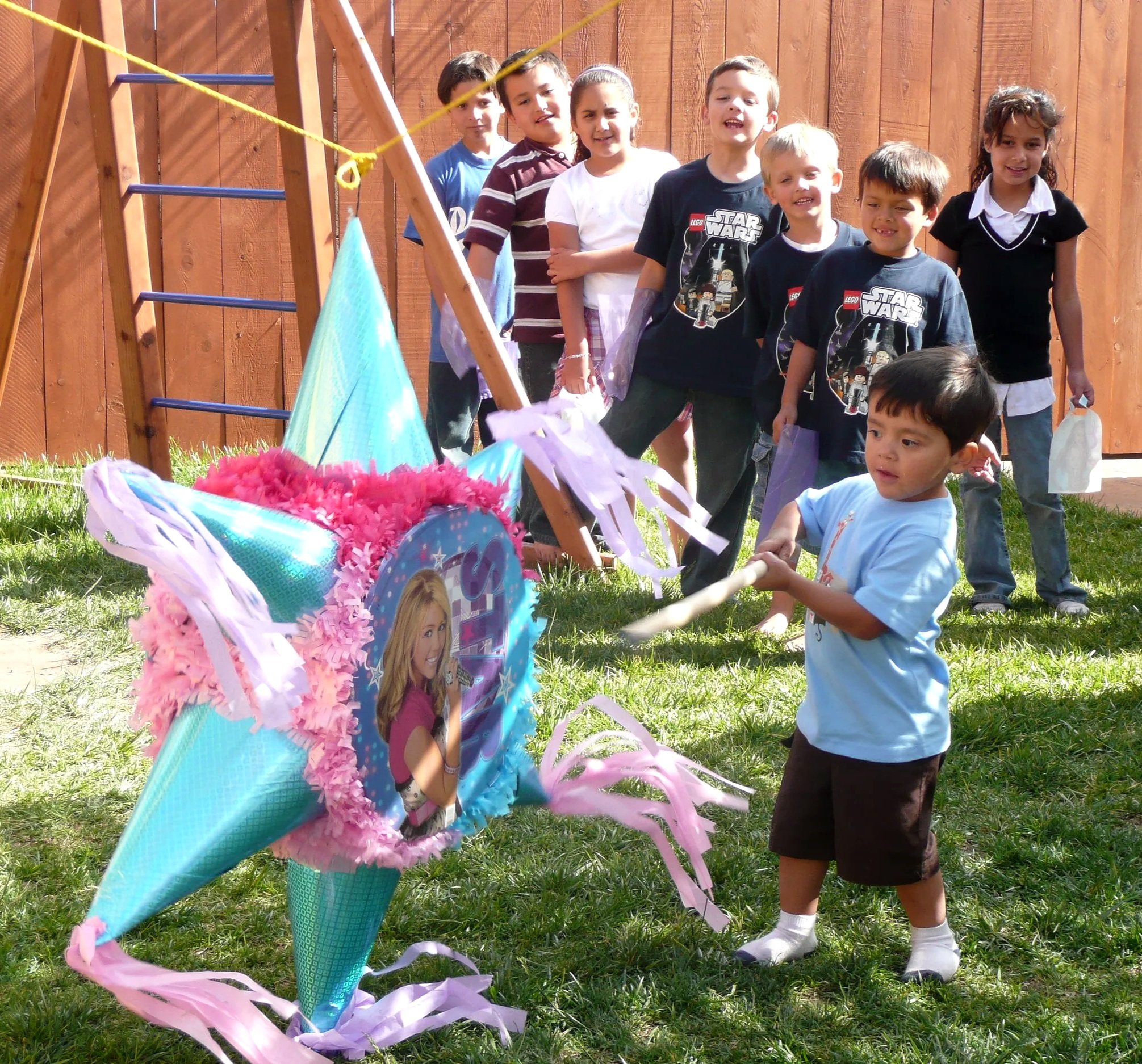 Children standing in line outdoors near a fence, waiting for a piñata to be broken during a celebration.