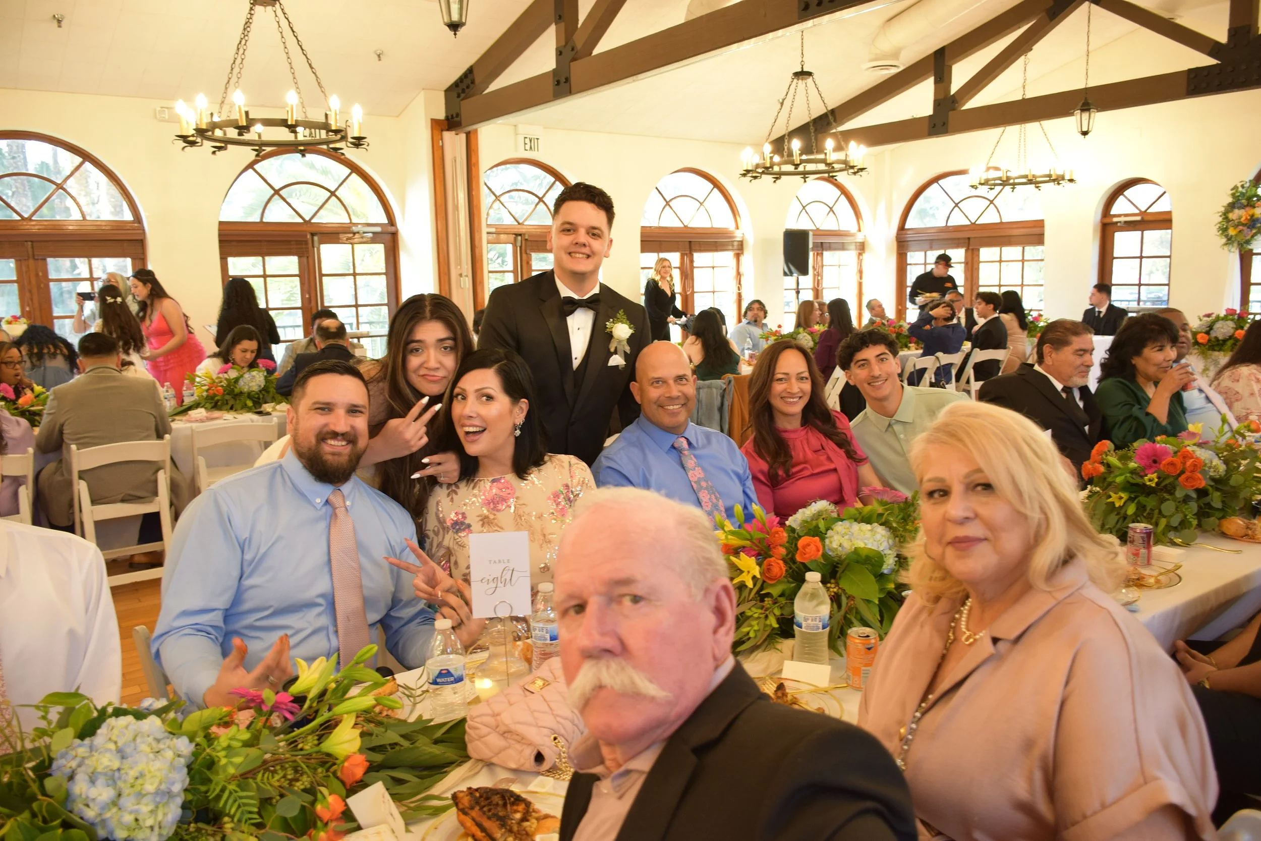 A group of people at a formal event, including a young man in a tuxedo and various adults sitting at decorated tables with flowers, in a well-lit banquet hall with large windows and chandeliers.