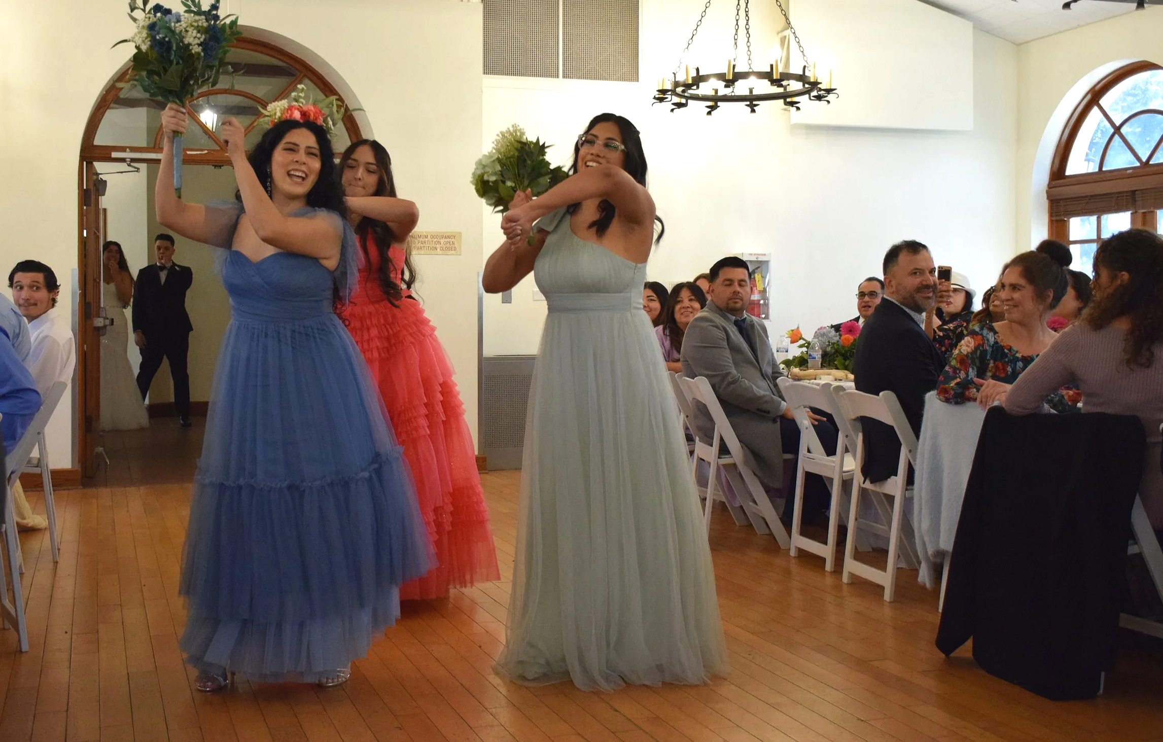 Three women dressed in colorful formal gowns standing in the middle of a reception hall, holding flowers and smiling. Guests seated at tables are watching them, and a chandelier hangs from the ceiling.