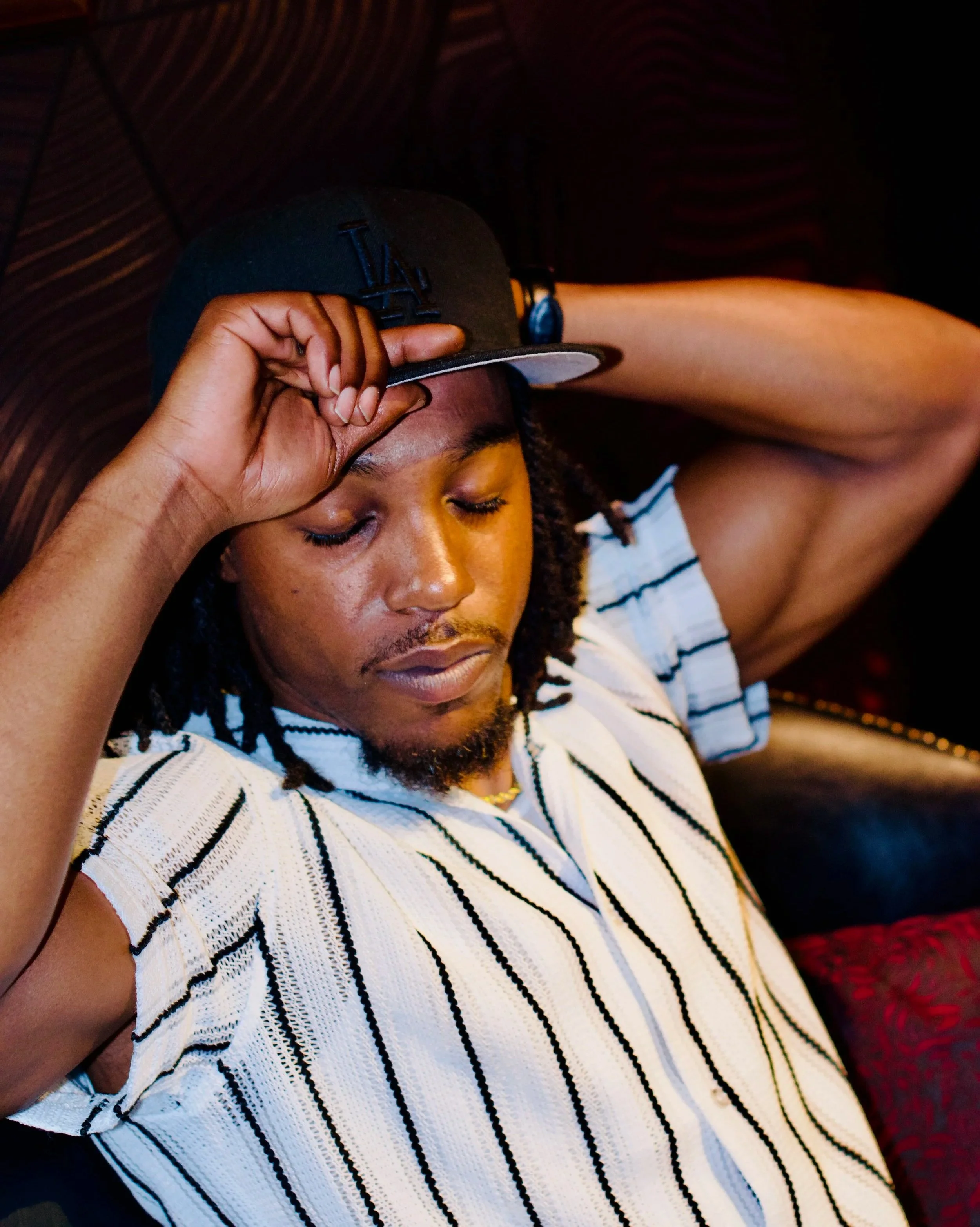 A person with curly hair wearing a black Los Angeles Dodgers baseball cap, a white and black striped shirt, with eyes closed, headphones on, and arm resting behind head.