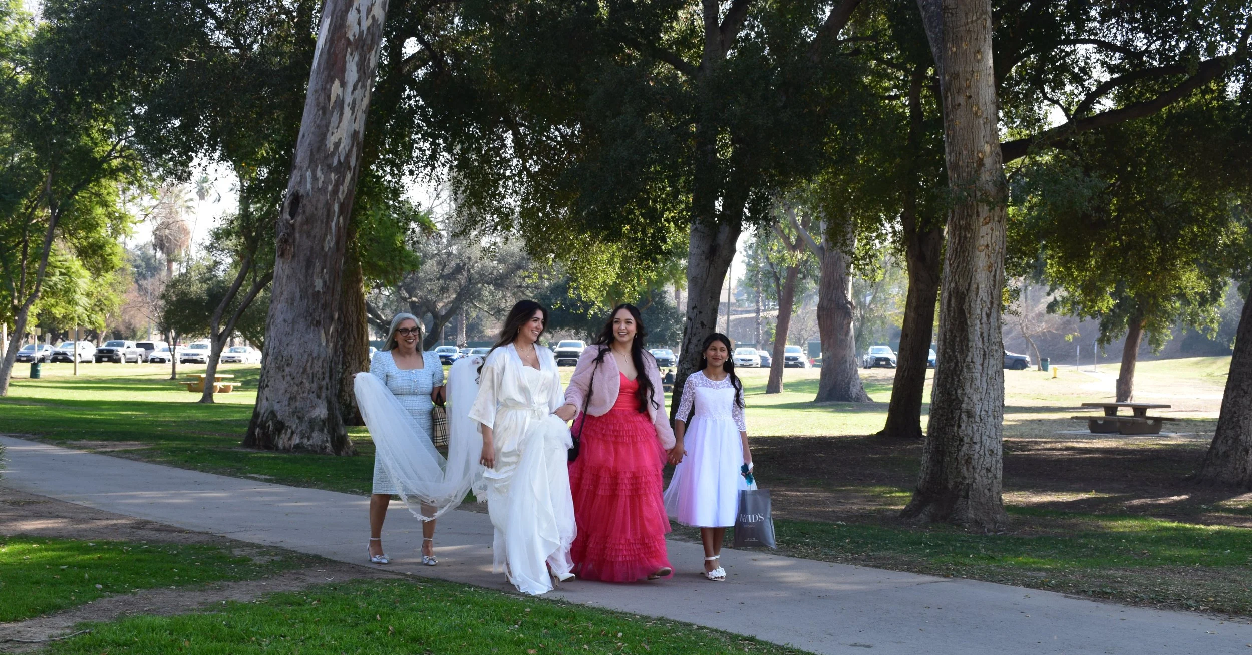 Group of five women in formal dresses walking in a park with trees and cars in the background.