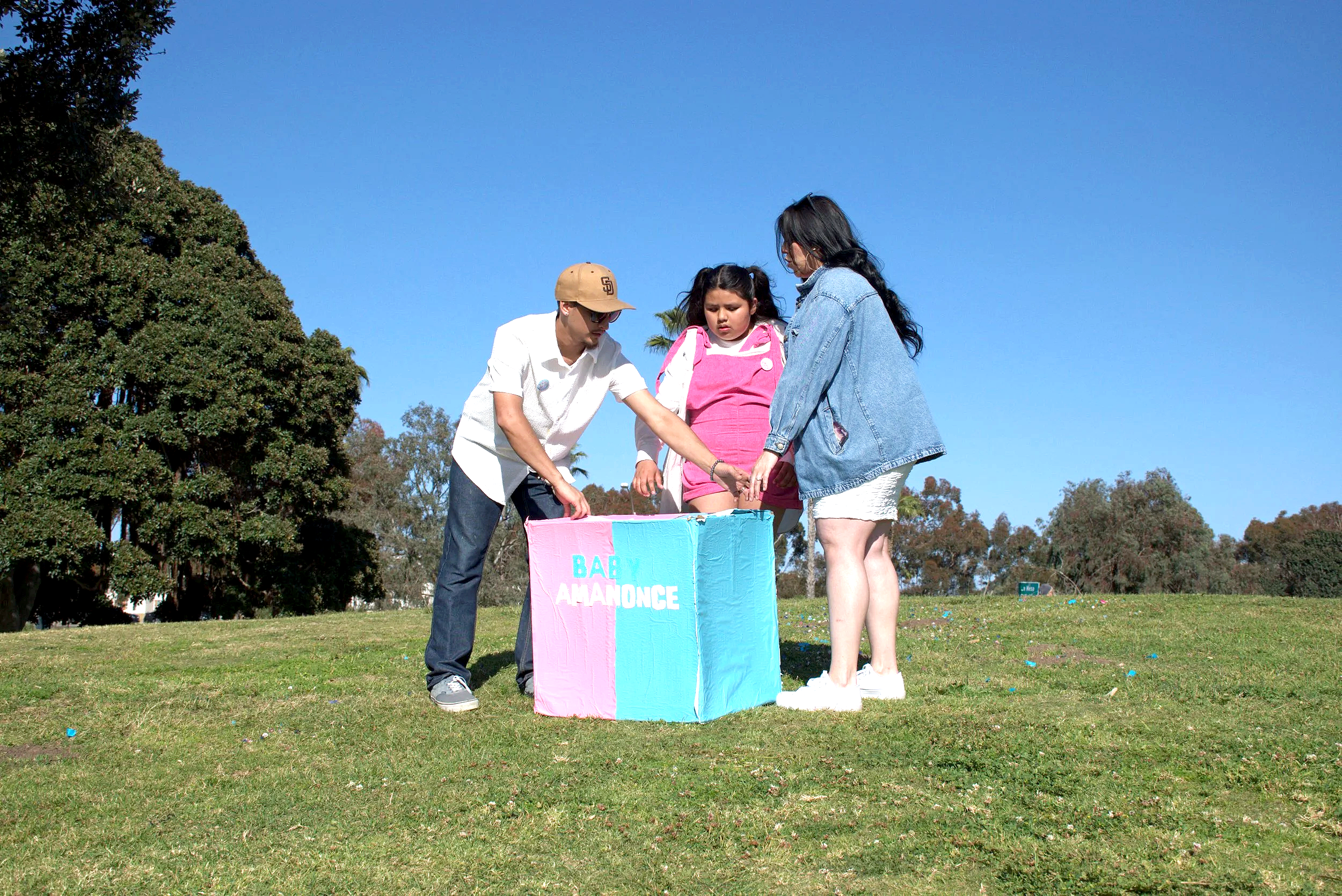 A family of three standing outdoors in a park on a sunny day near a pink and blue sign that reads "Baby Announcement" with trees and a clear blue sky in the background.