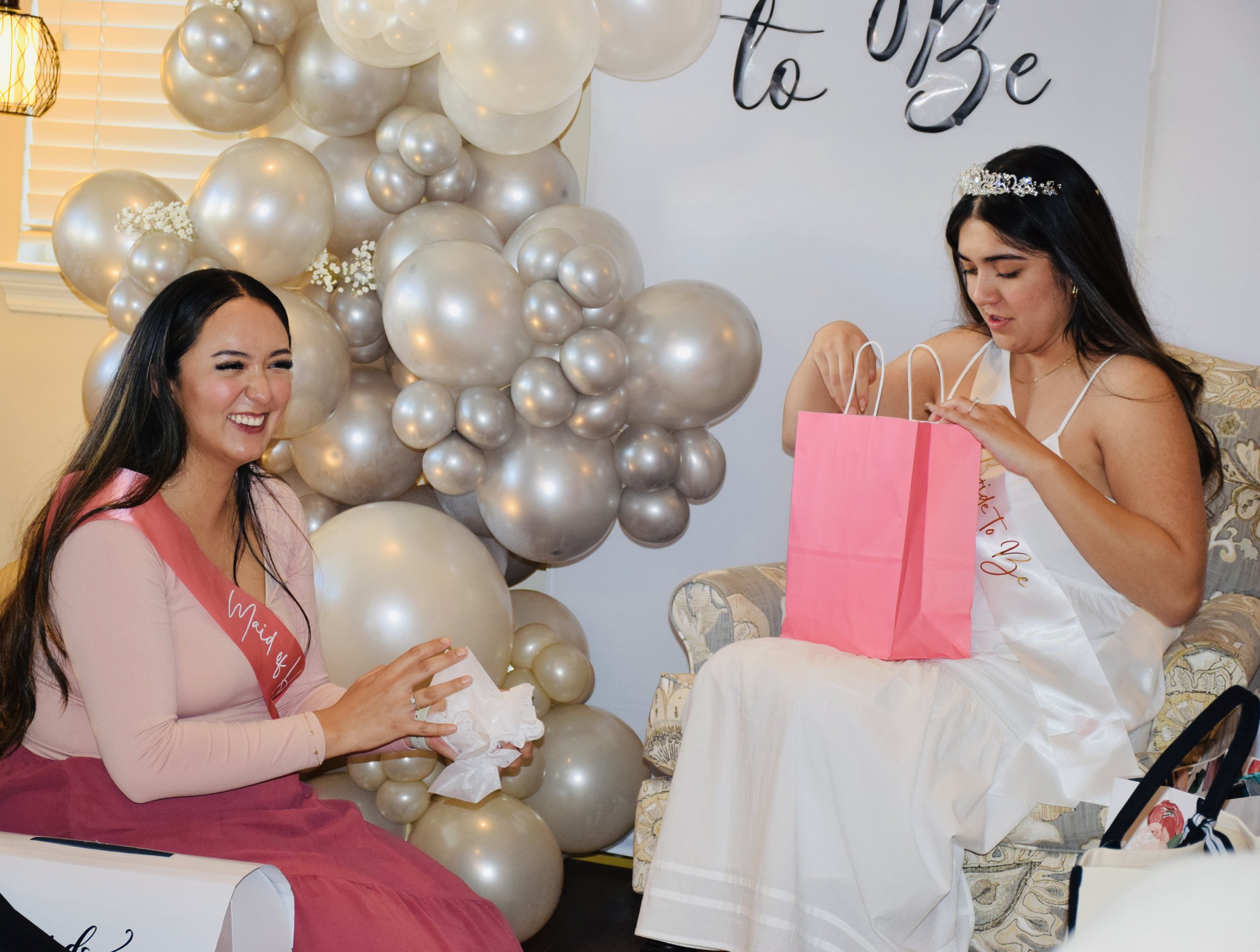 Two women at a bridal shower, one wearing a pink dress and a sash that reads 'Maid of Honor', the other wearing a white dress and a tiara, sitting in a room decorated with a large arrangement of gold and white balloons and a sign that says 'To Be' in the background, with a pink gift bag on her lap.