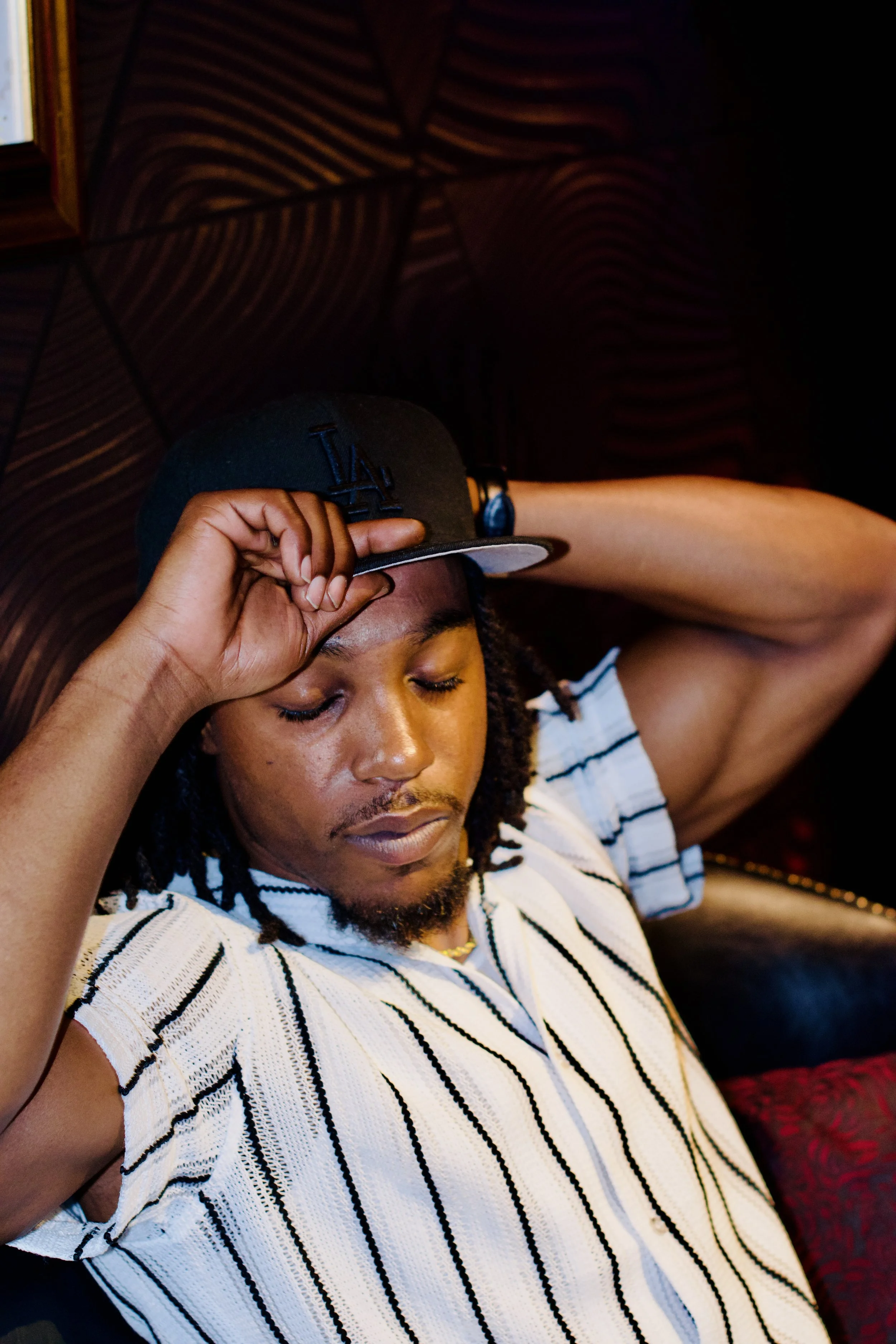 A young man with dreadlocks and a beard, wearing a striped white and black shirt and a black LA baseball cap, relaxing with eyes closed and hand resting on forehead, sitting on a dark patterned couch in a dimly lit room.