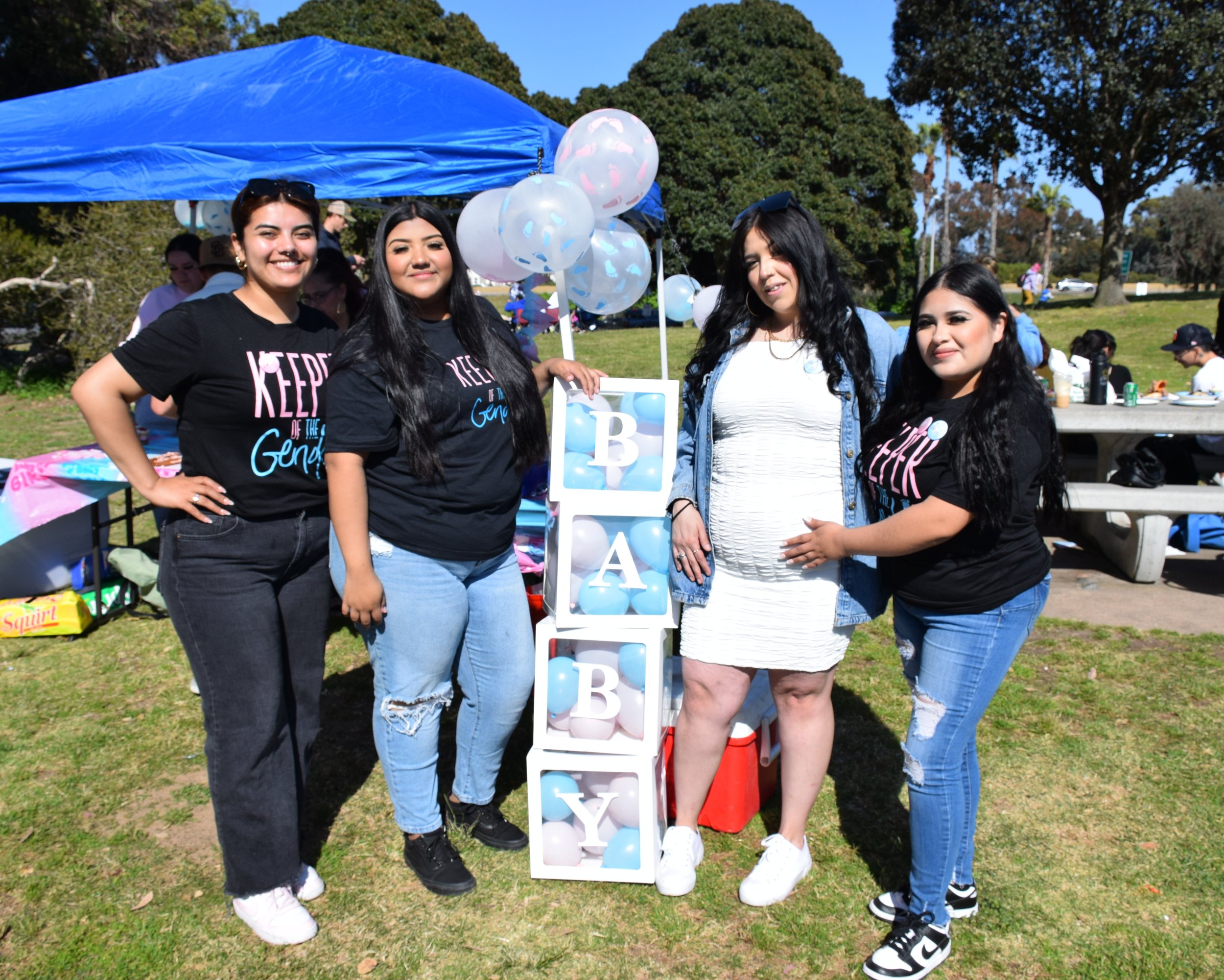 Four young women standing together outdoors at a daytime event, with a blue canopy, balloons, and a vertical arrangement of blocks spelling 'GIRL' in the background.