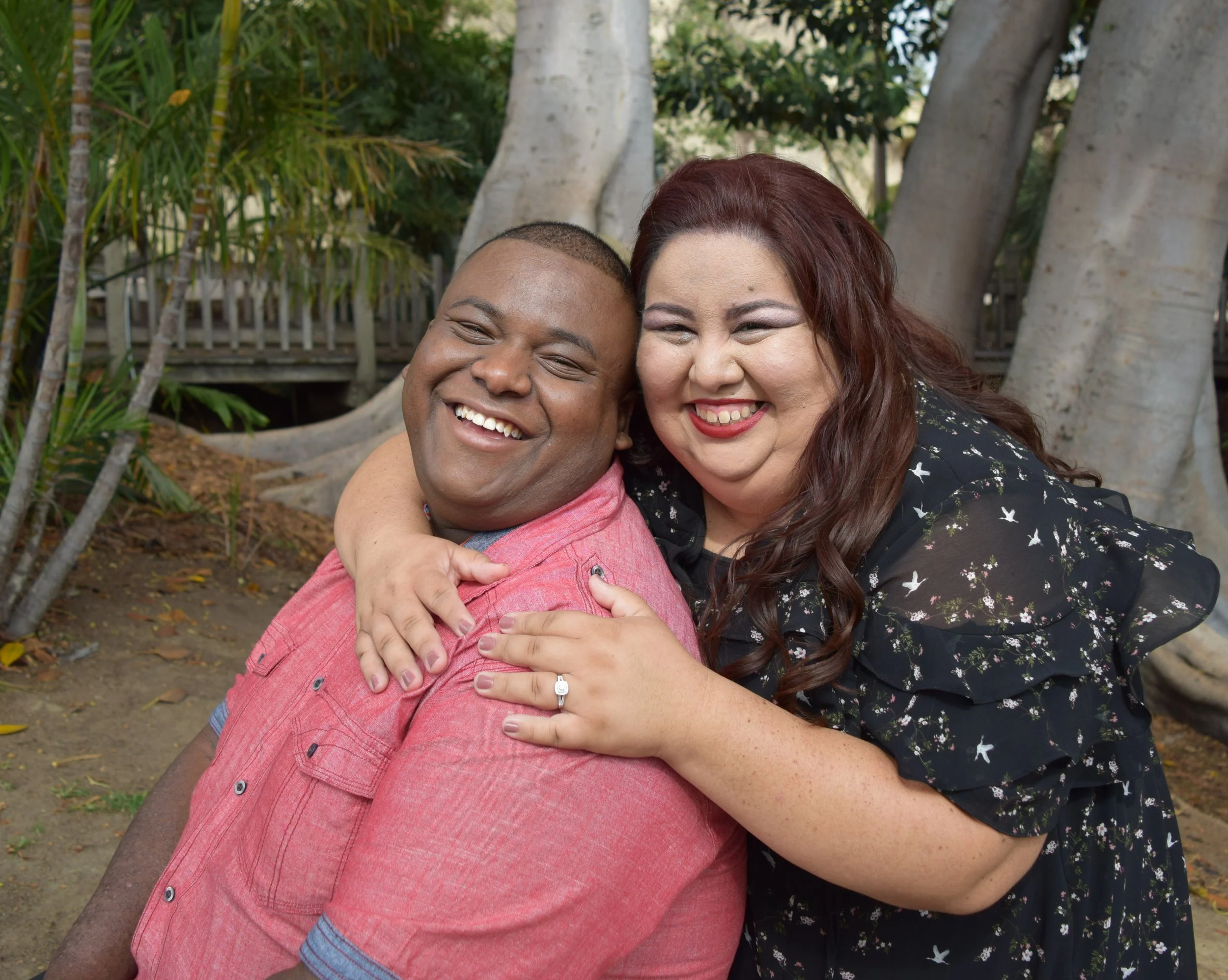 A happy man and woman embracing outdoors during daytime, surrounded by trees and nature, smiling widely.