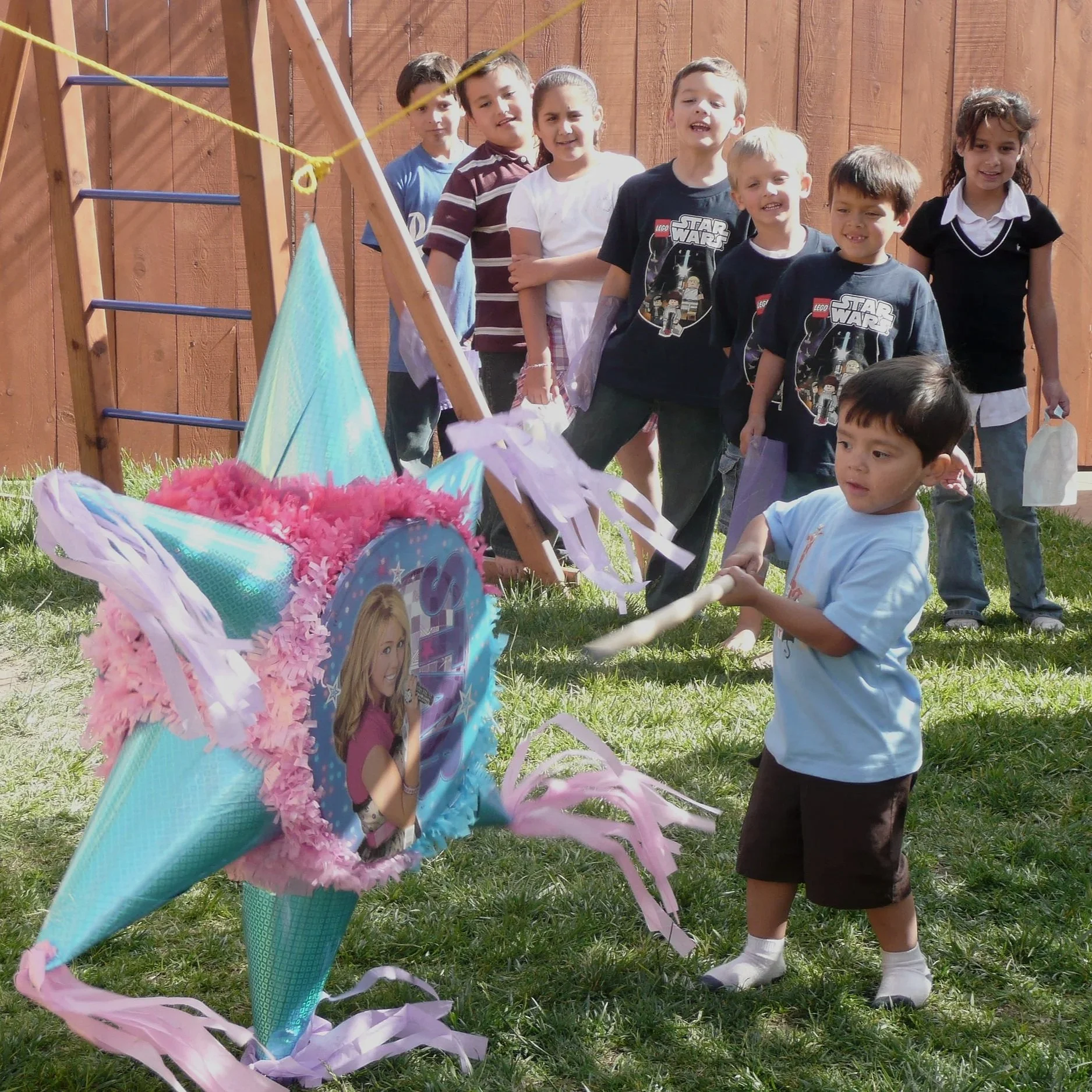 Children at a birthday party stand in line outdoors near a fence, with a colorful piñata in the foreground.
