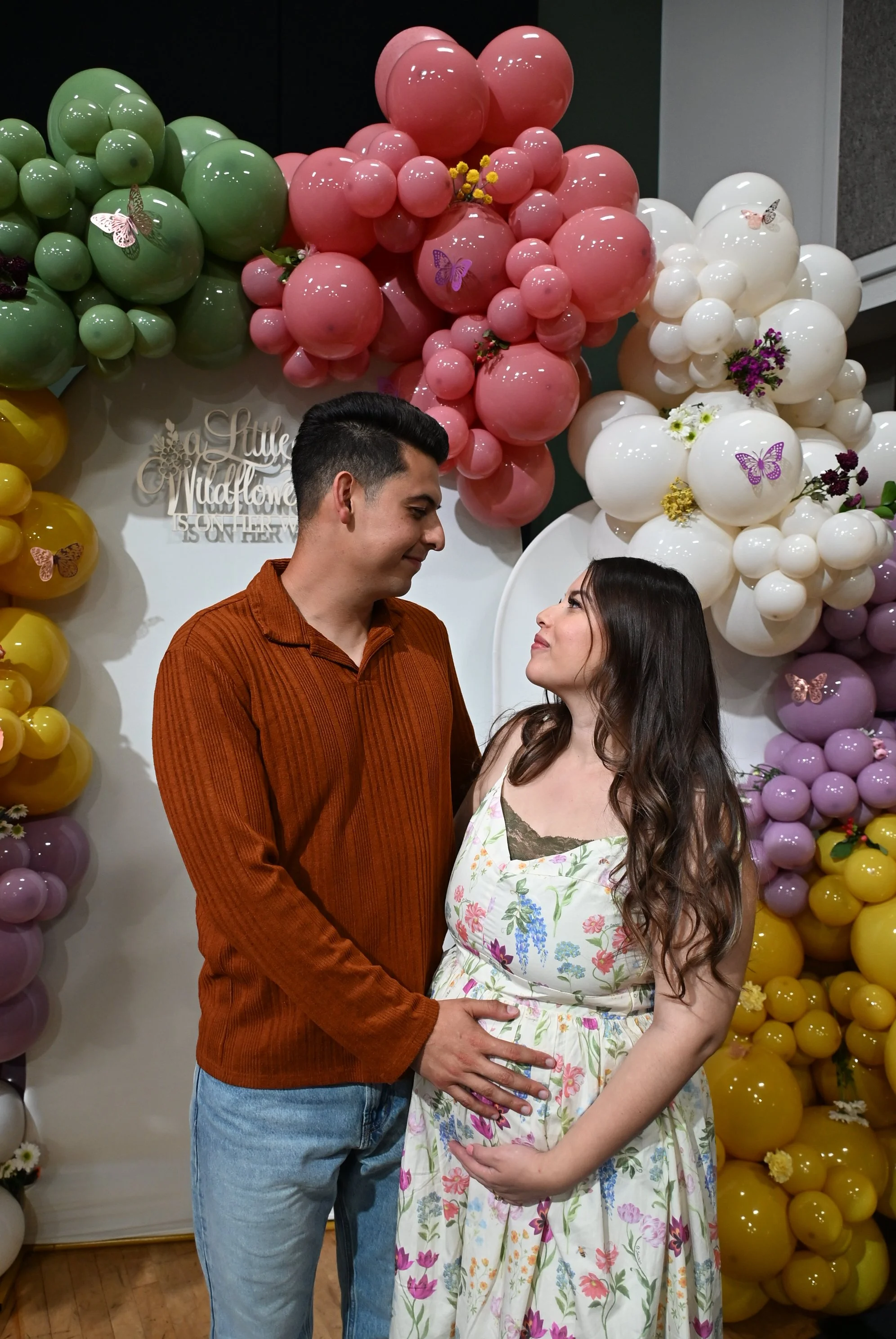 A couple is standing in front of a colorful balloon arrangement for a gender reveal celebration. The man is wearing a rust-colored shirt and jeans, and the woman is wearing a floral dress and appears pregnant.
