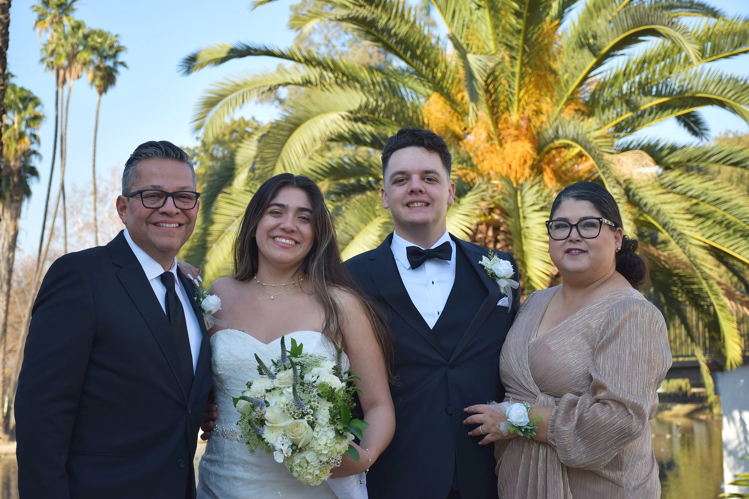 Group of four people dressed in formal attire, standing outdoors in front of palm trees, smiling at the camera during a sunny day.