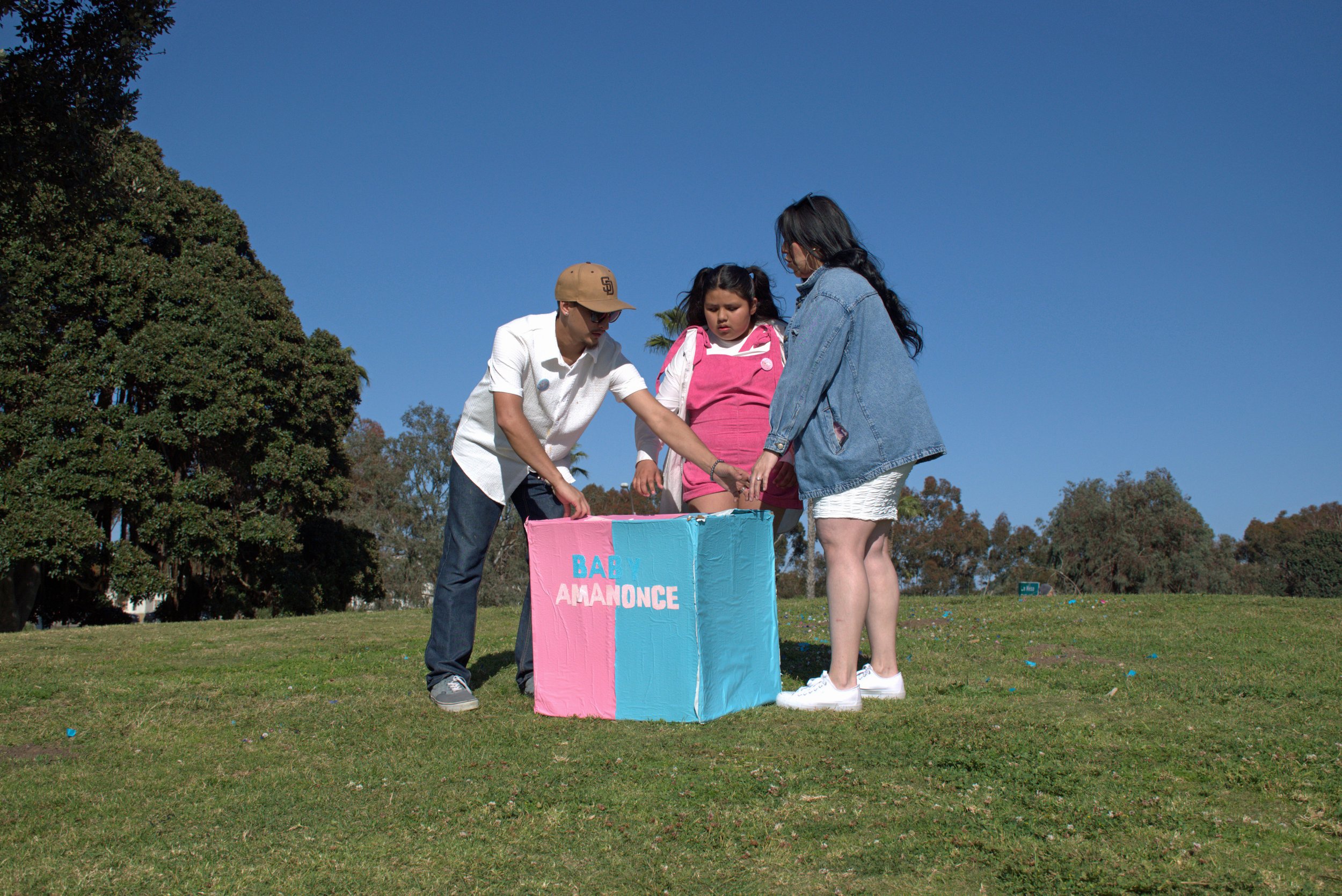 A family of three, a man, a woman, and a young girl, are standing outside in a park during daytime, gathered around a pink and blue booth or box with a sign that reads 'BABY AMONCE'. The man is wearing a tan cap, sunglasses, a white shirt, and dark j