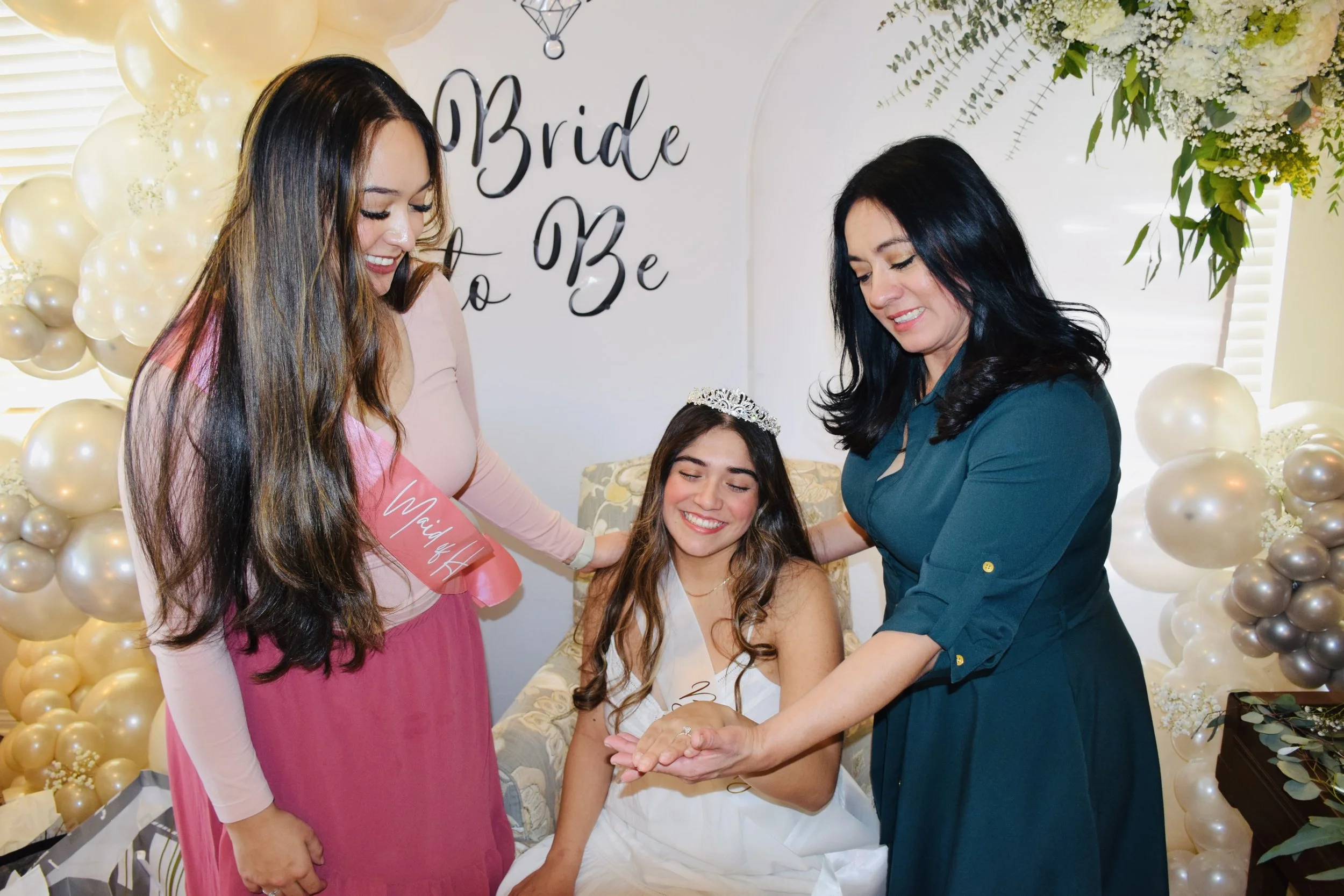 A bride sitting in a decorated room, surrounded by women, with one woman placing a ring on her finger. The room has balloon decorations and a sign that reads 'Bride to Be'.