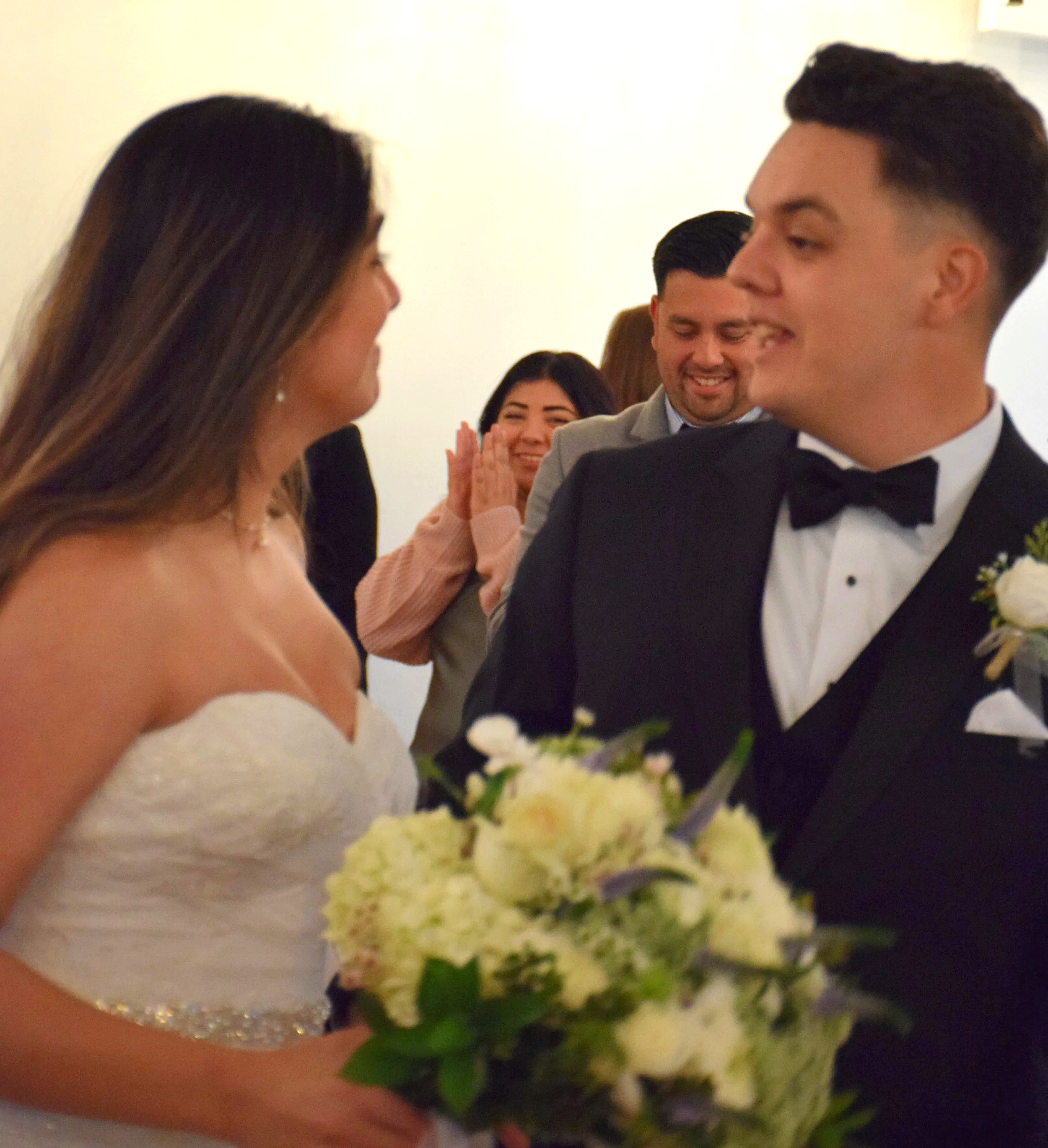 A wedding scene with a bride and groom smiling at each other, surrounded by guests, in a well-lit indoor setting.