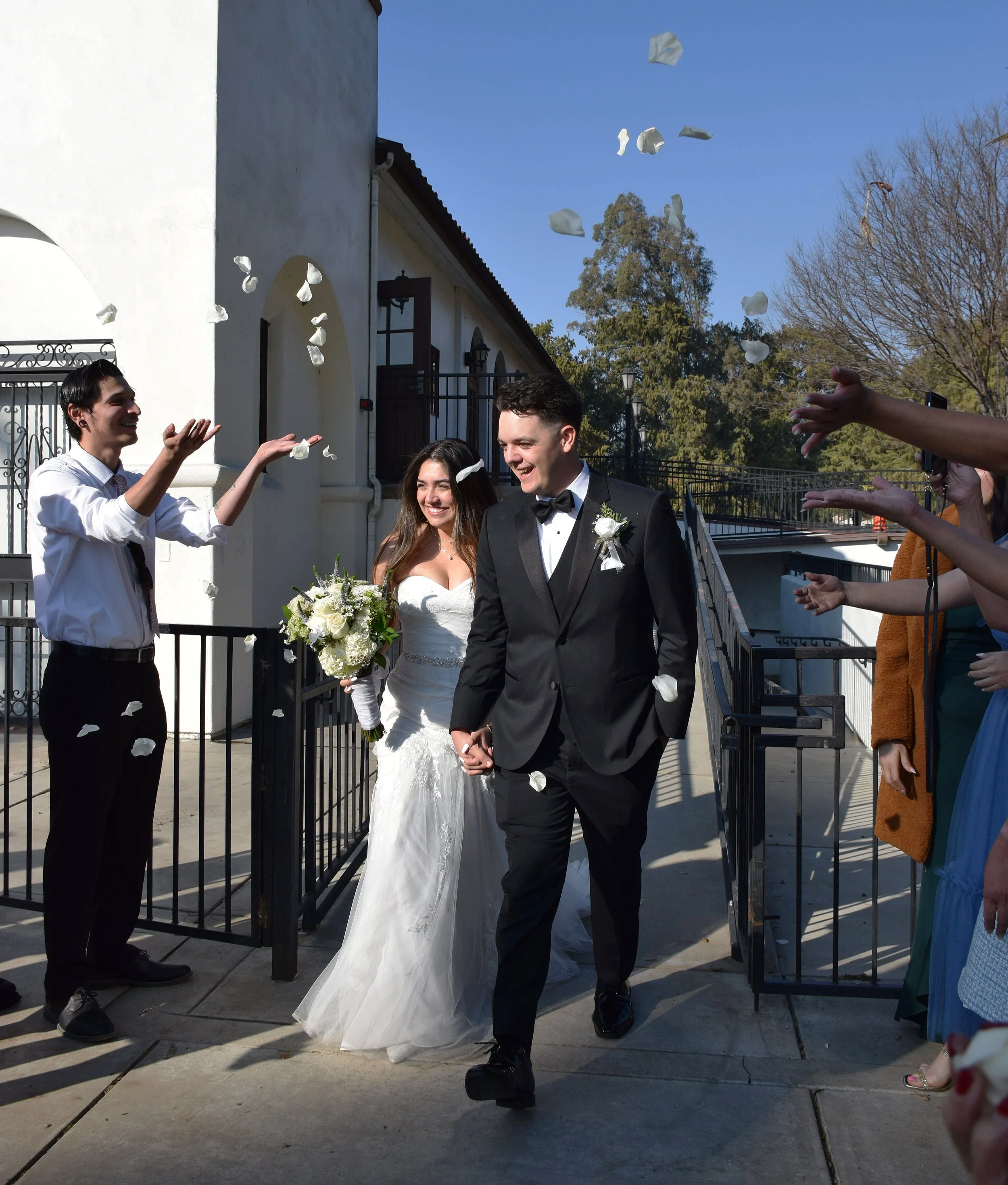A bride and groom hold hands and walk out of a building, smiling, after a wedding ceremony, with guests throwing flower petals in the air on a sunny day.