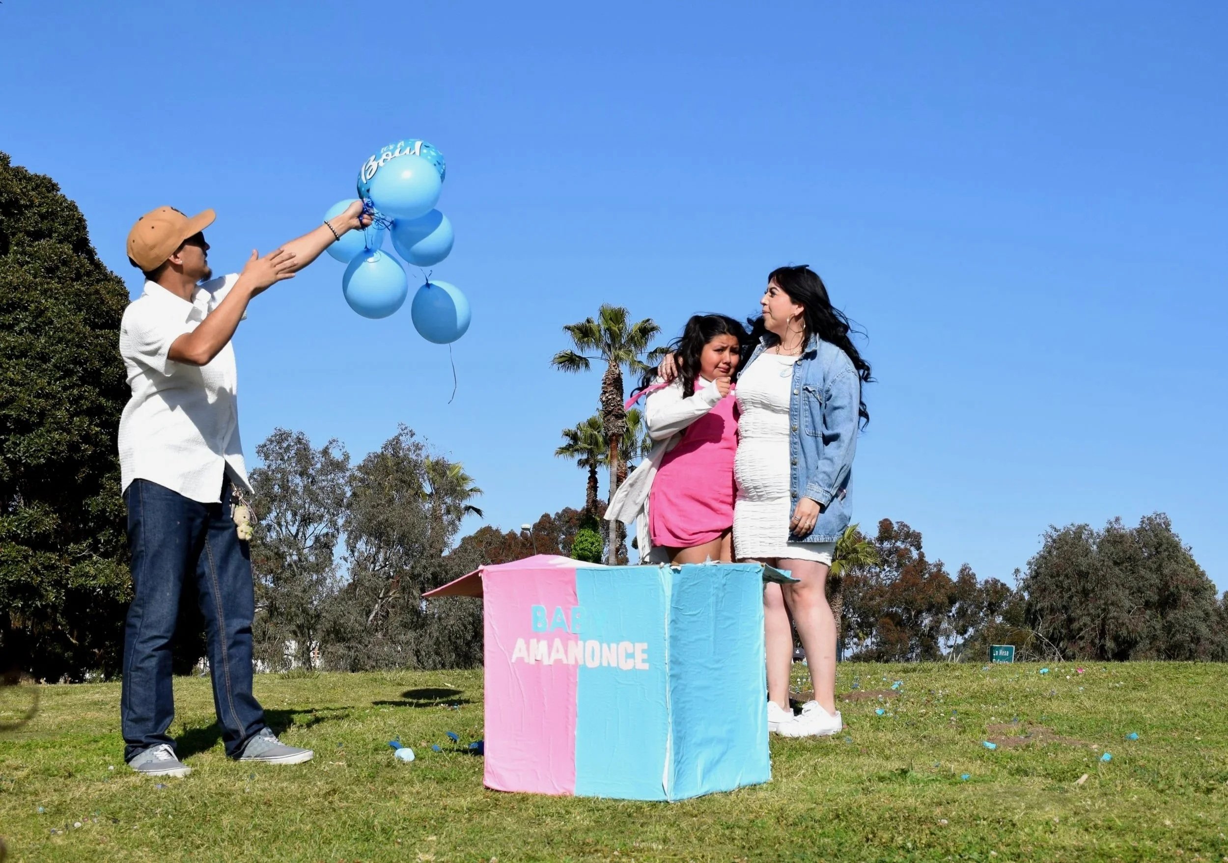 A woman dressed in white with a denim jacket and another woman in a pink dress are standing on a grassy field with trees and palm trees in the background, surrounded by blue balloons being handed over by a man in a white shirt, sunglasses, and a beige cap during daytime.