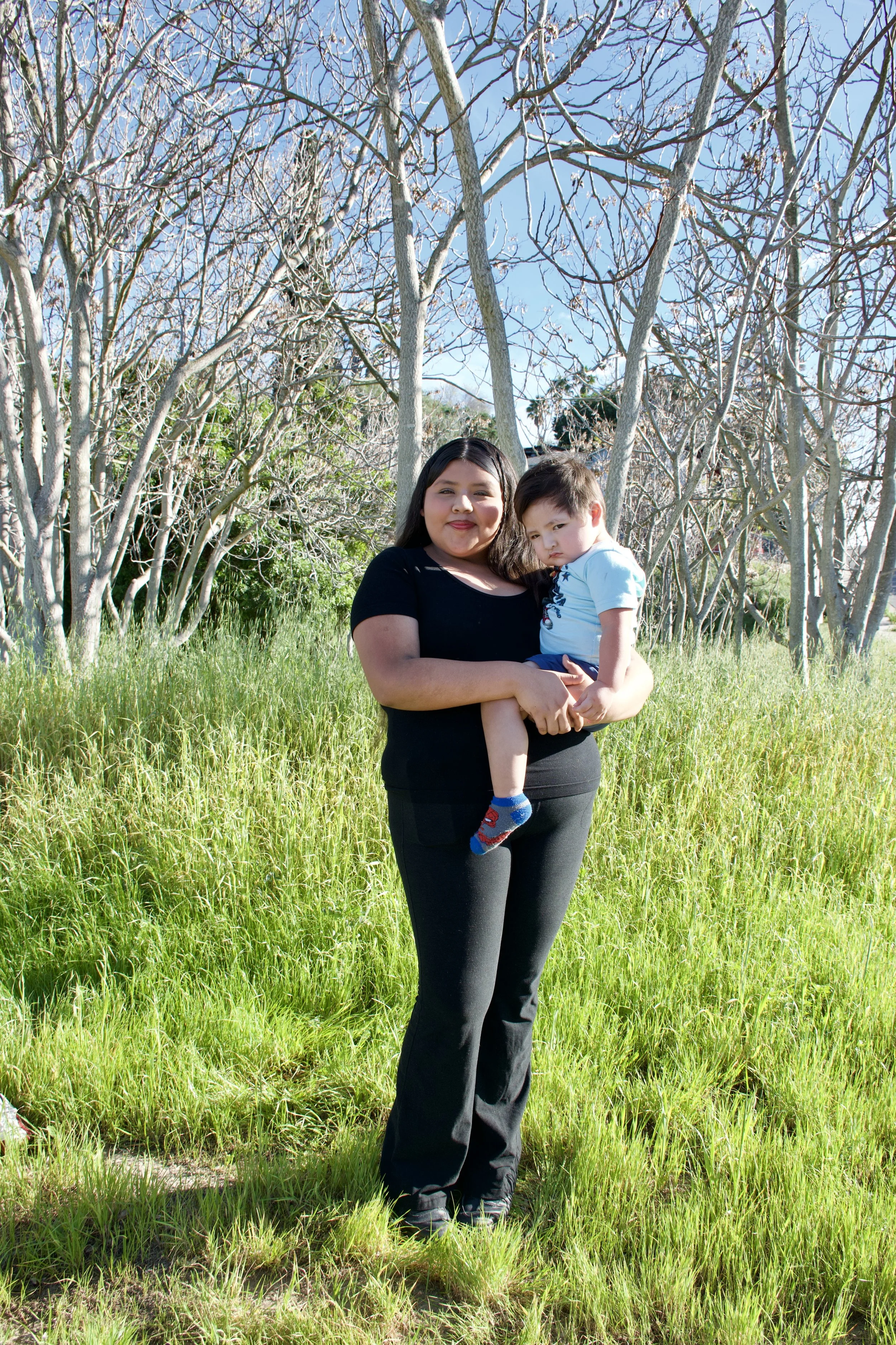 A woman holding a young boy in a grassy area with bare trees and a clear blue sky in the background.