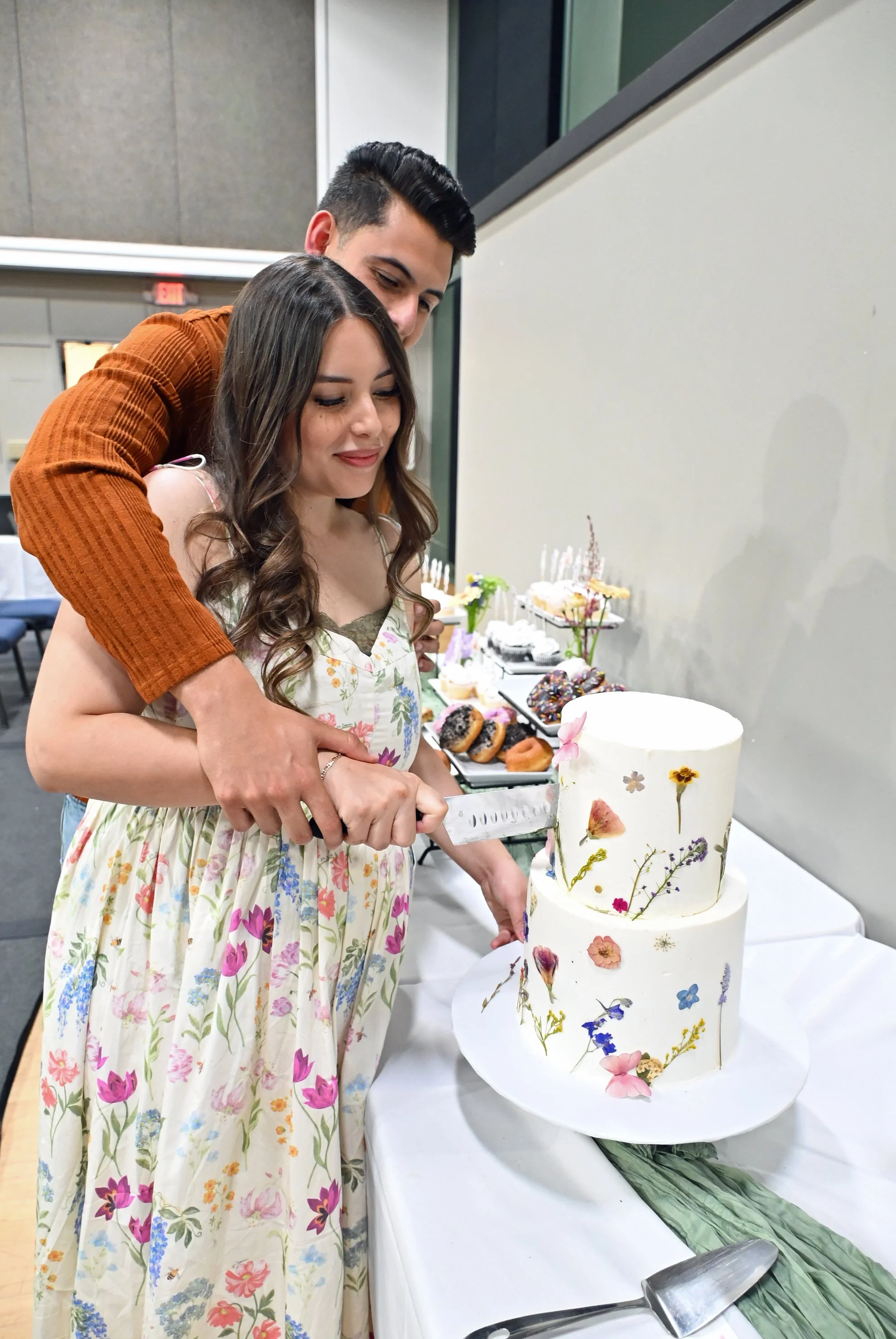 A woman in a floral dress with another person, both are cutting a two-tiered white cake decorated with pressed flowers. The cake is placed on a table draped in white cloth, with desserts and a fork nearby.